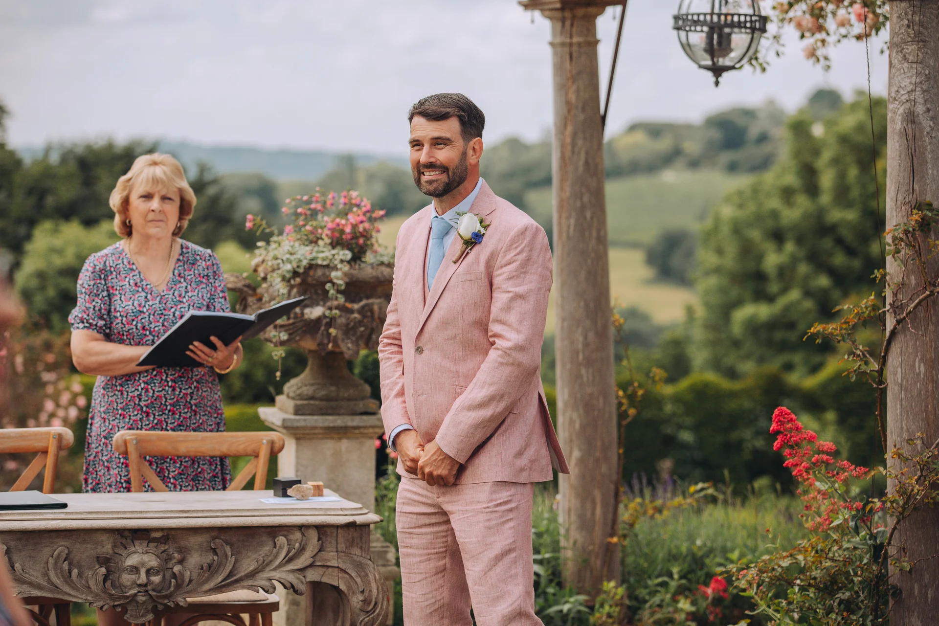A smiling groom wearing a pink suit and boutonniere stands at an outdoor altar during a scenic celebrant-led ceremony with rolling hills in the background.