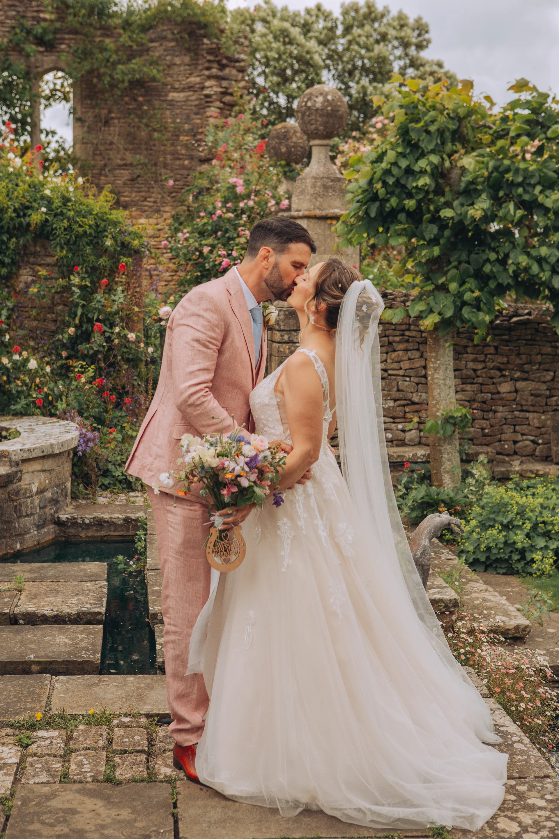 A bride and groom share a tender kiss in a romantic historic walled garden, surrounded by stone ruins, vibrant climbing roses, and lush greenery.