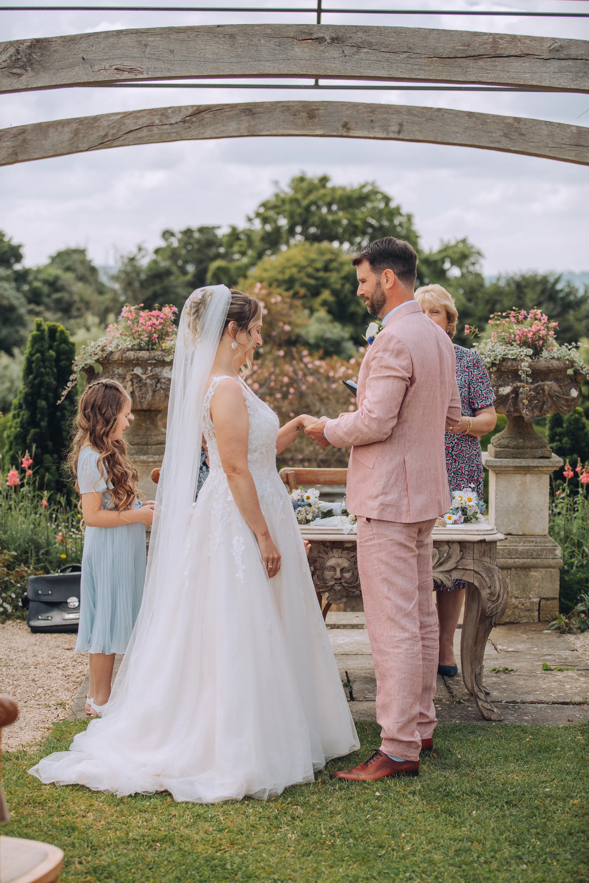 An outdoor wedding ceremony taking place in a garden where the bride and groom exchange rings near a rustic stone table under an arched wooden arbor.