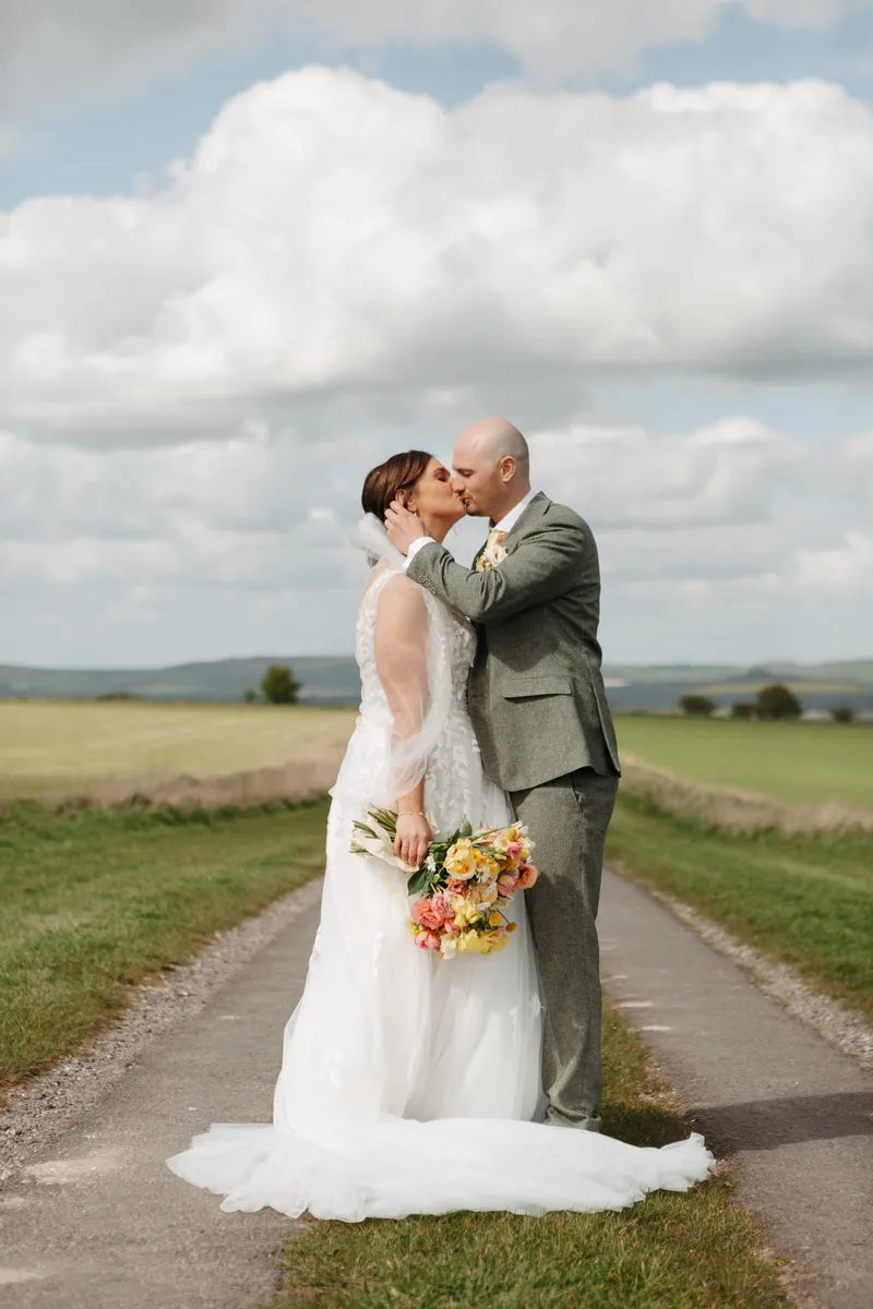 Newlyweds share a tender kiss on a country path surrounded by rolling green hills under a dramatic cloudy sky.