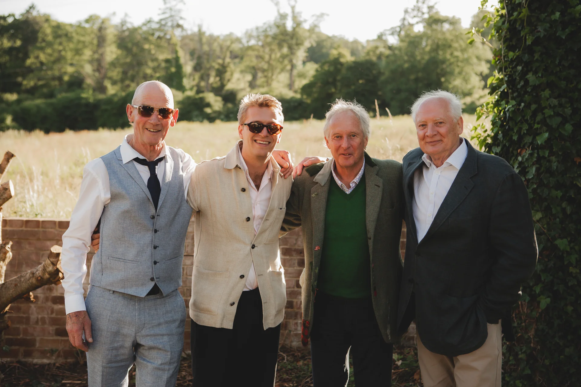 Four male wedding guests stand outdoors in a sunny garden setting, smiling together with arms around each other.