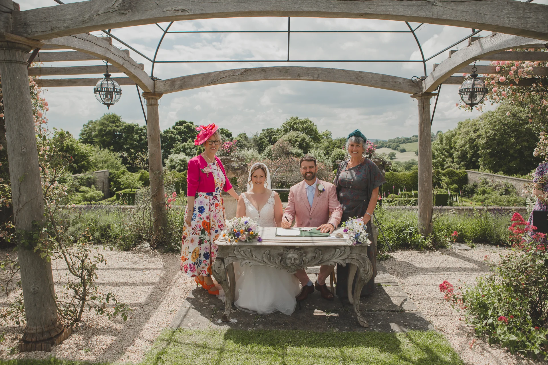 The bride and groom smile while signing their marriage register beneath a wooden pergola in a lush country garden, flanked by family members in wedding attire.