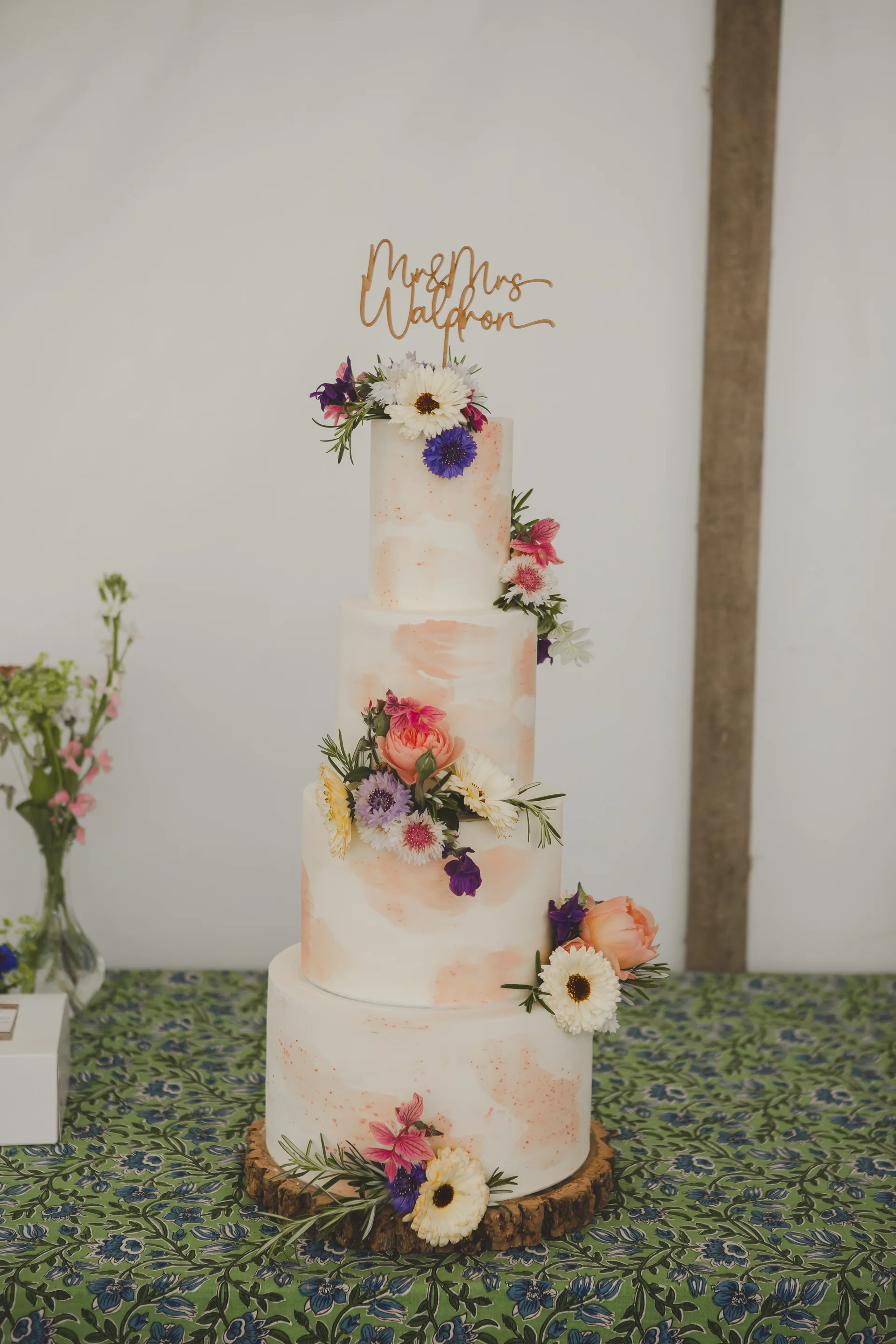 A stunning four-tier wedding cake featuring watercolour brushstrokes and fresh wildflowers rests on a rustic wooden stand atop a patterned green tablecloth.