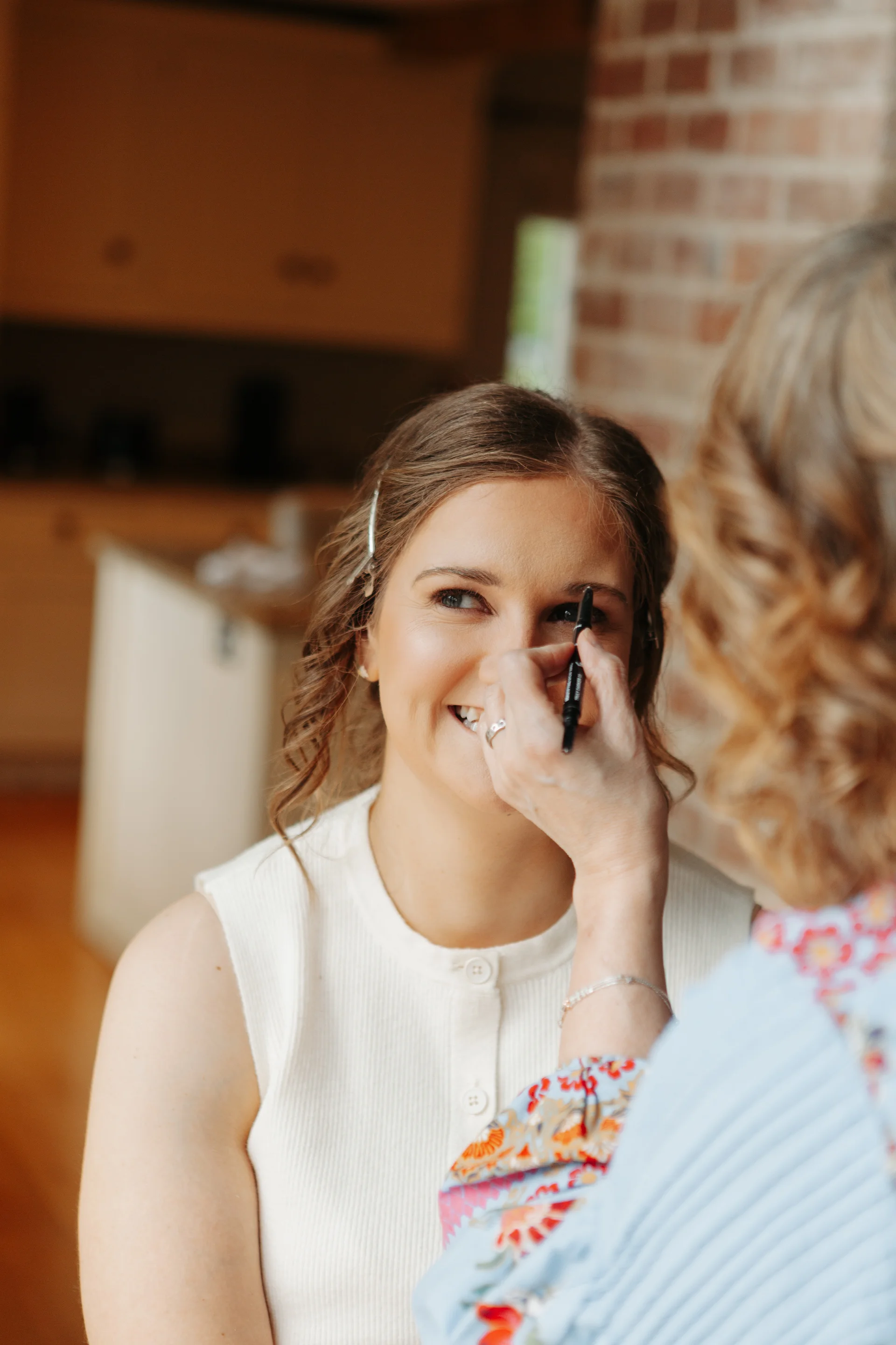 A smiling bride receives eyebrow makeup application from a stylist during morning bridal preparations indoors.
