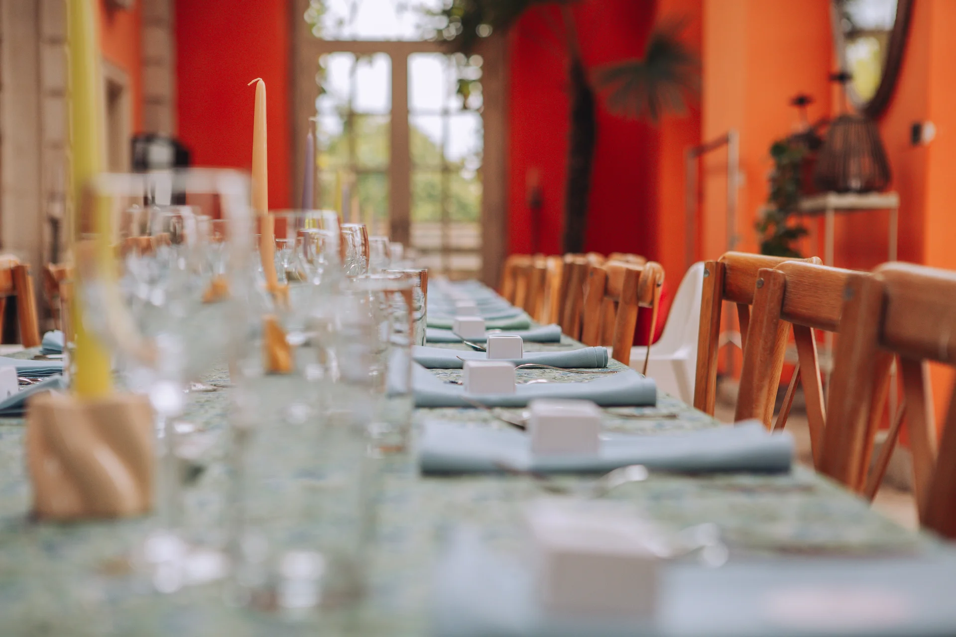 A long banquet wedding table is beautifully set with pale blue napkins, small favour boxes, and tall taper candles against a vibrant orange room.