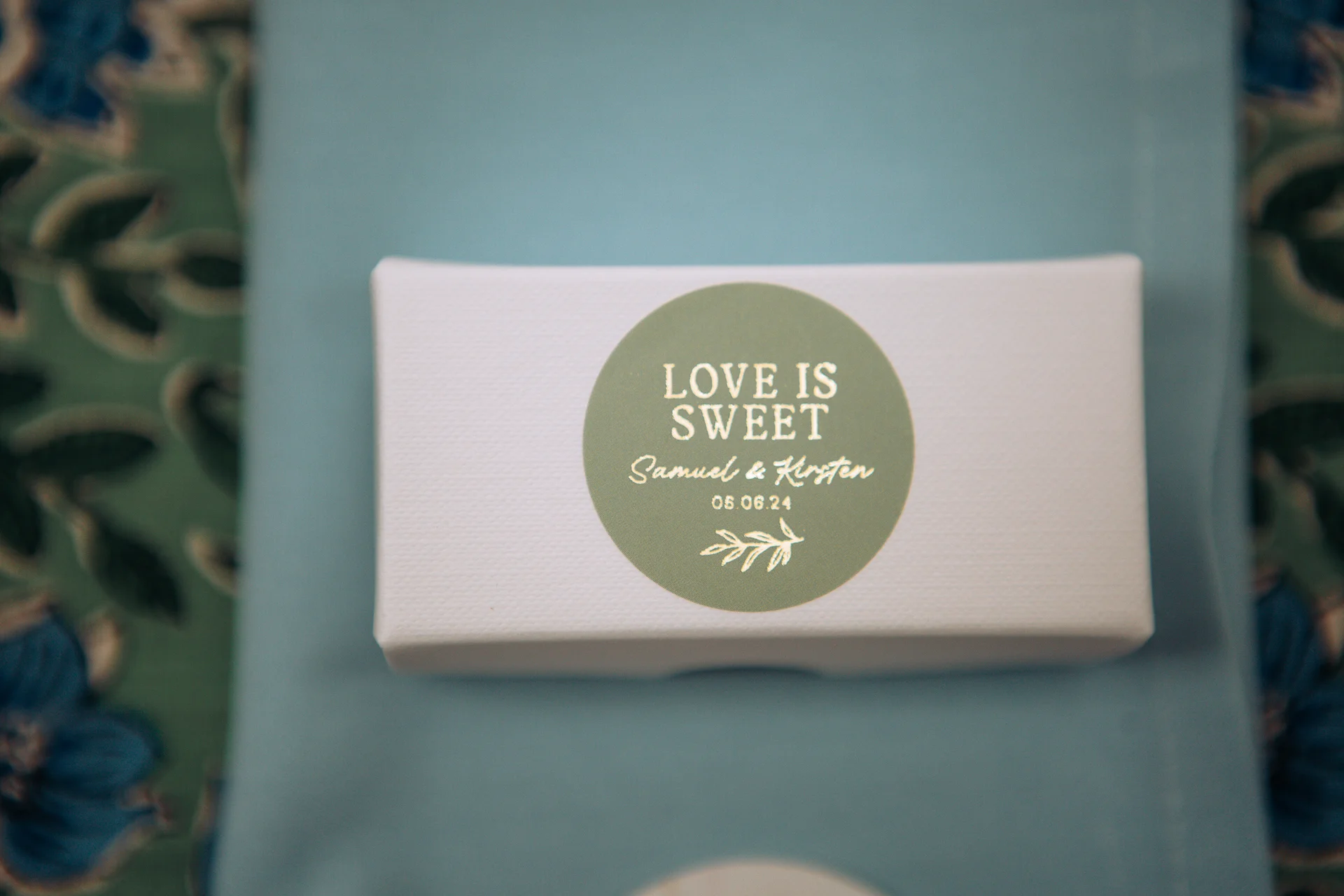 A close-up shot of a white wedding favour box featuring a round sage green sticker with gold foil lettering that reads love is sweet alongside names and a date.