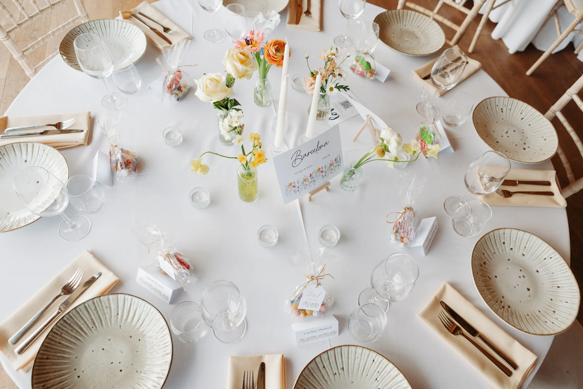 An overhead view of a beautifully set round wedding table featuring neutral linens, textured ceramic plates, gold cutlery, delicate bud vases with spring florals, and personalized place settings.