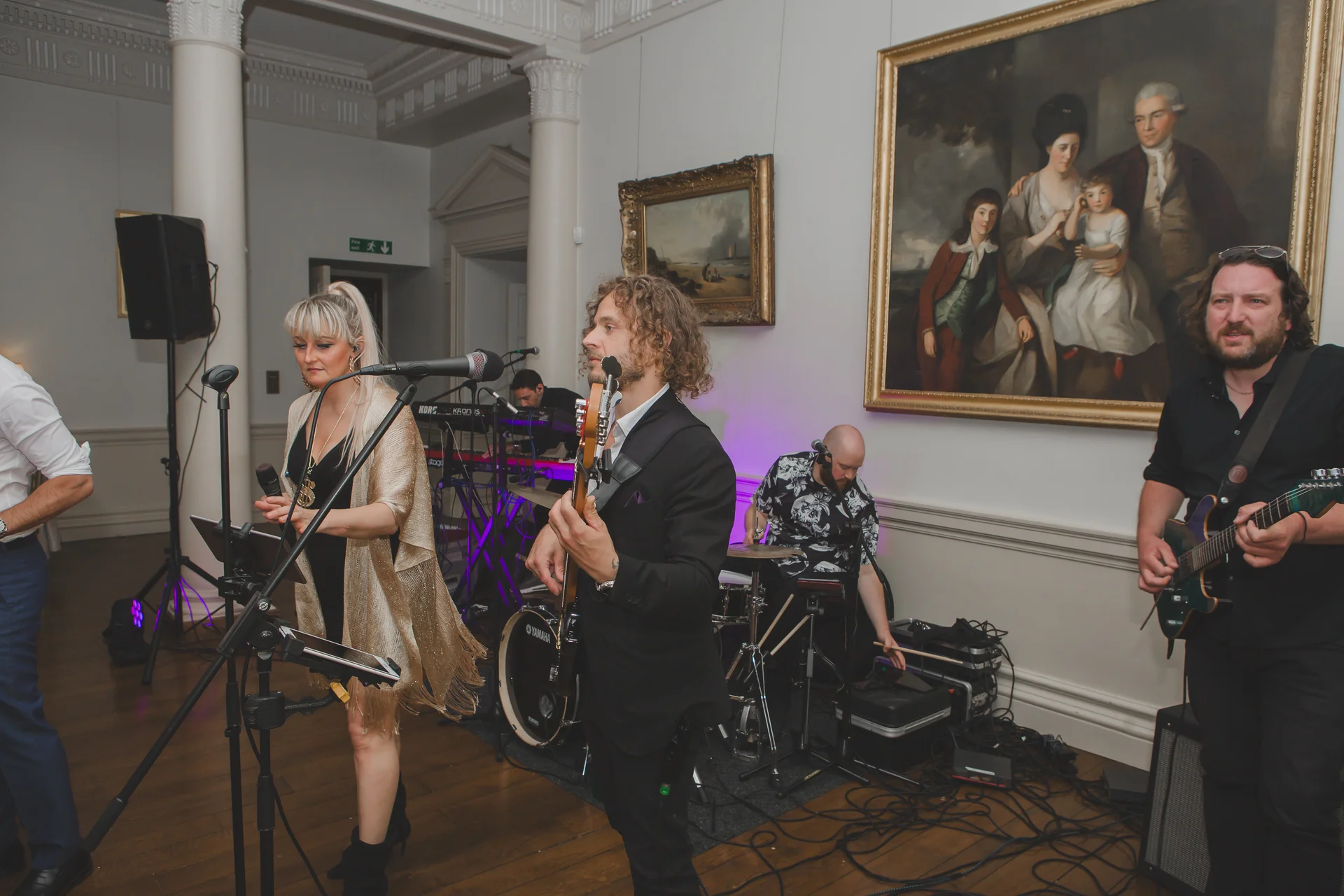 A wedding function band performs live in a stately home interior featuring ornate oil paintings on the walls and a wooden floor.