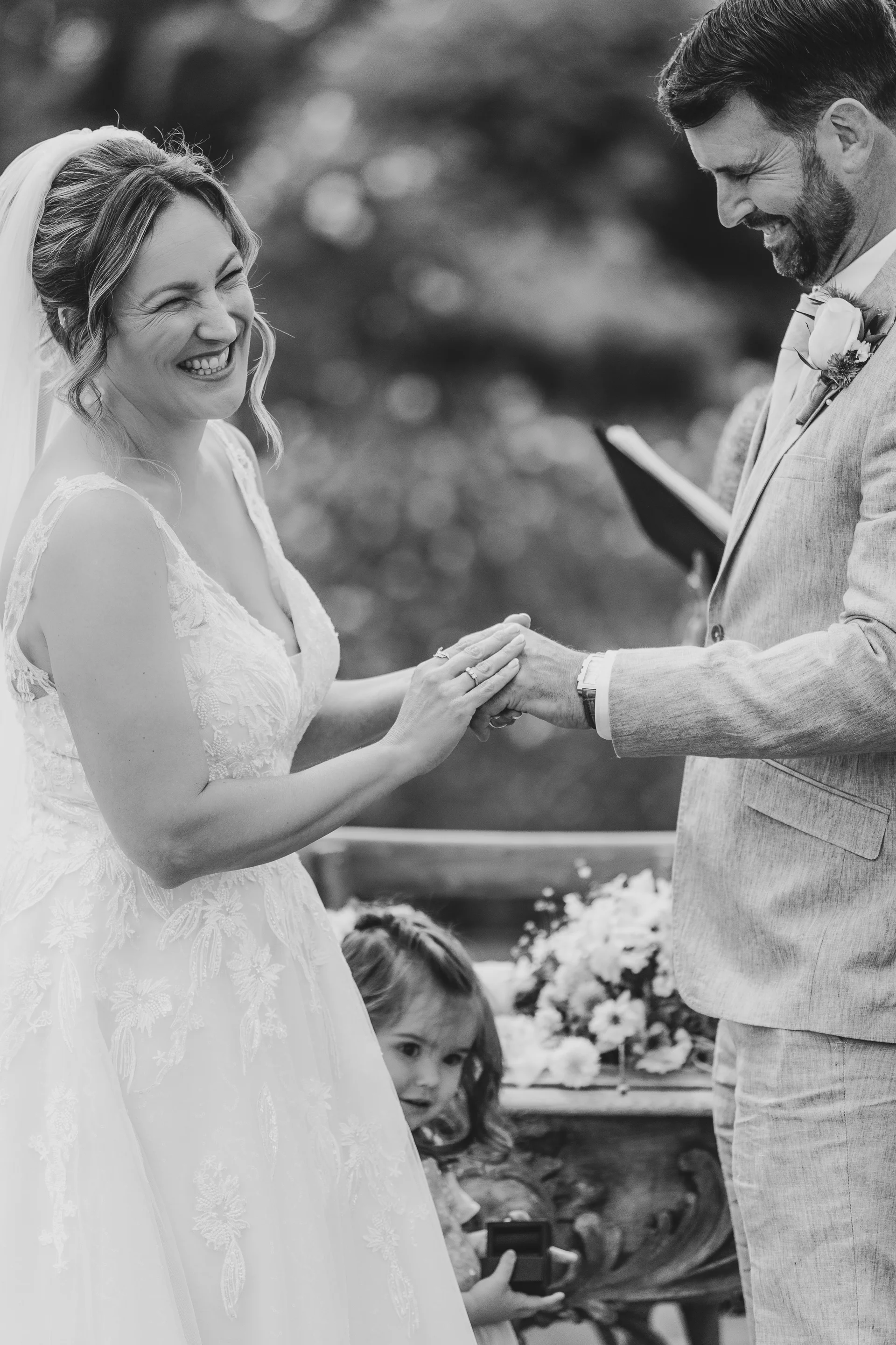 A joyful bride and groom laugh as they exchange rings during their outdoor wedding ceremony, with a young child peeking out below them.