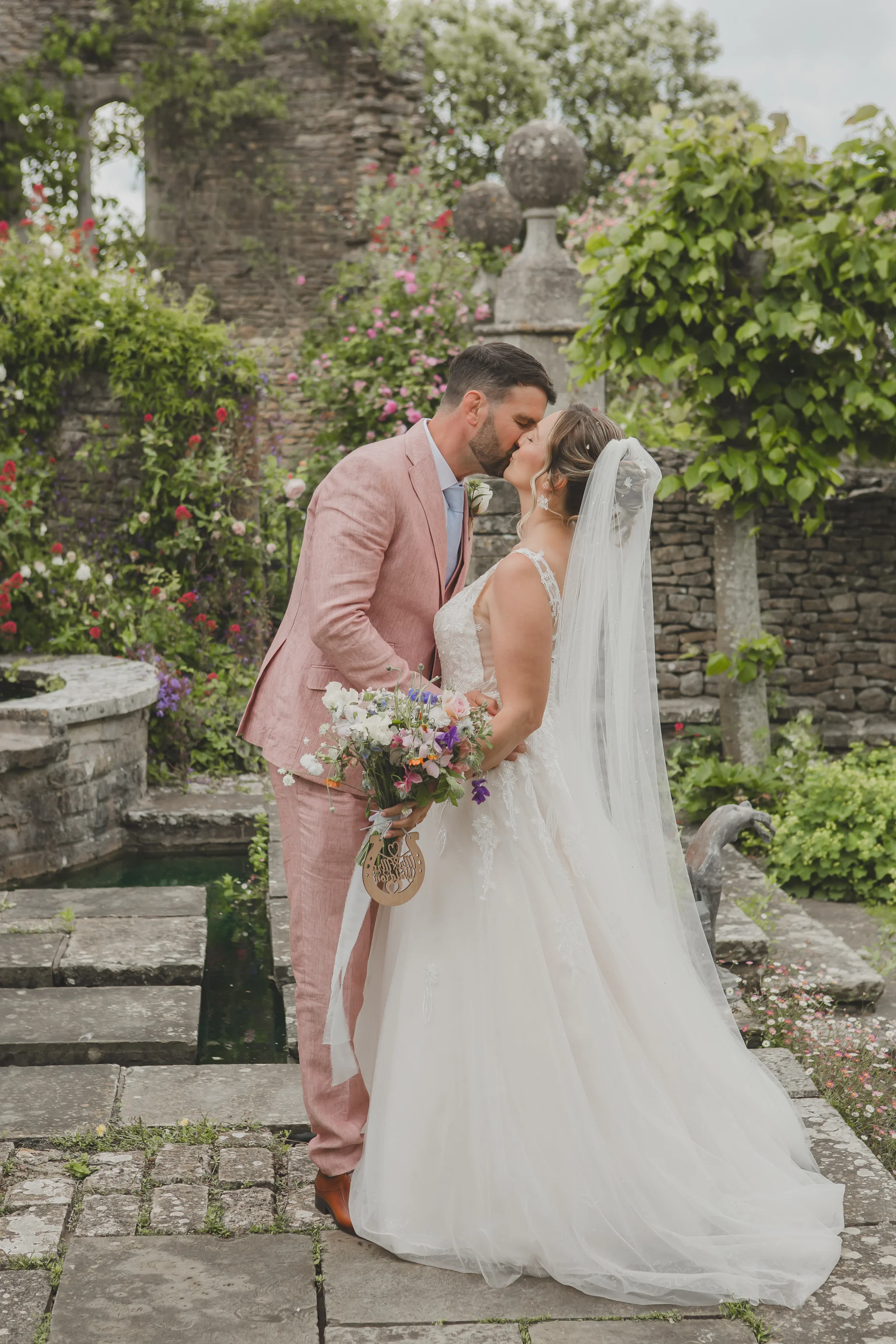 Newlyweds share a tender kiss in a picturesque walled garden featuring stone pathways, ancient ruins, and lush floral arrangements.