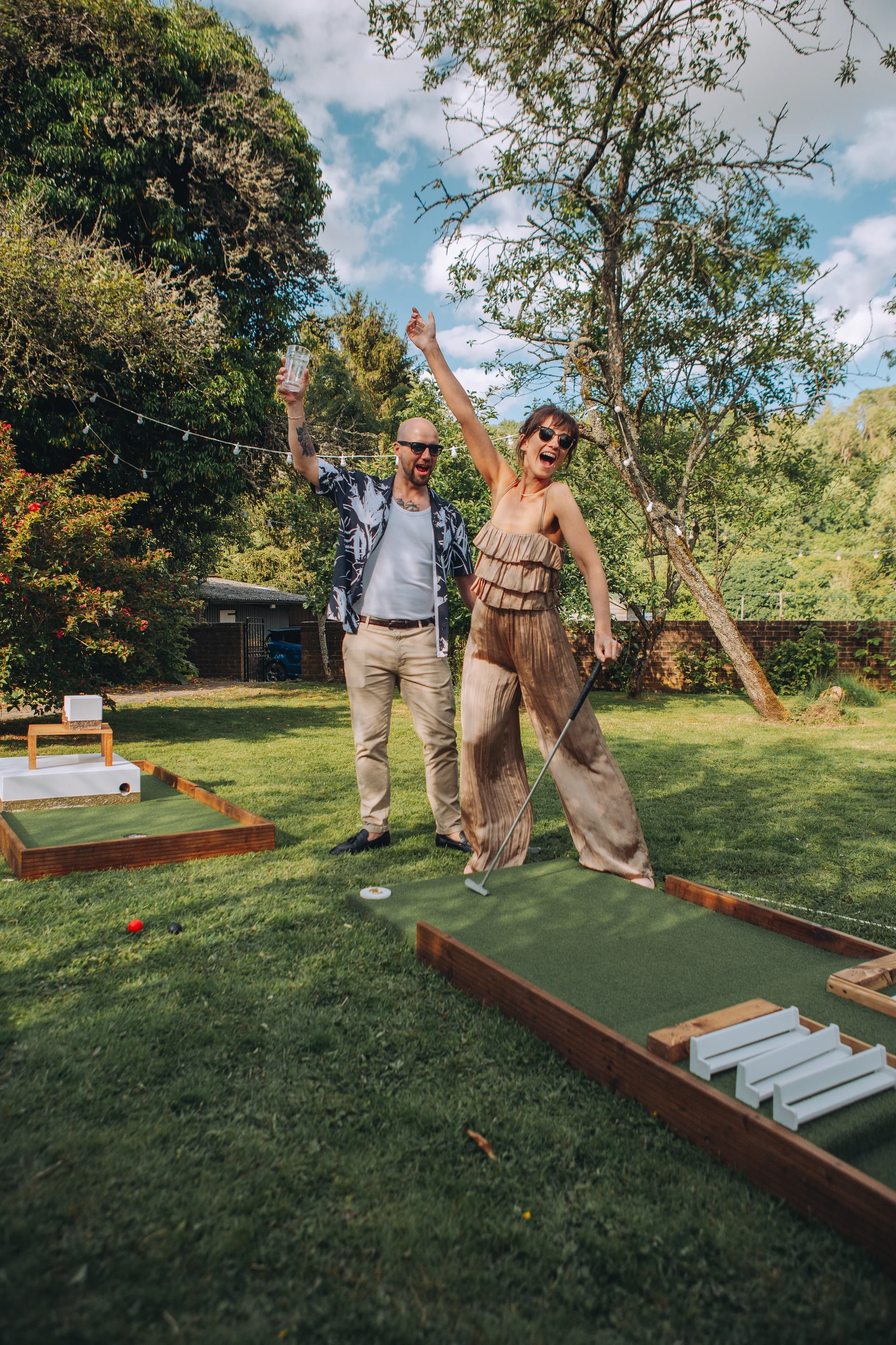 A joyful couple in casual attire play a game of mini golf on the lawn during an outdoor wedding reception in the sunshine.