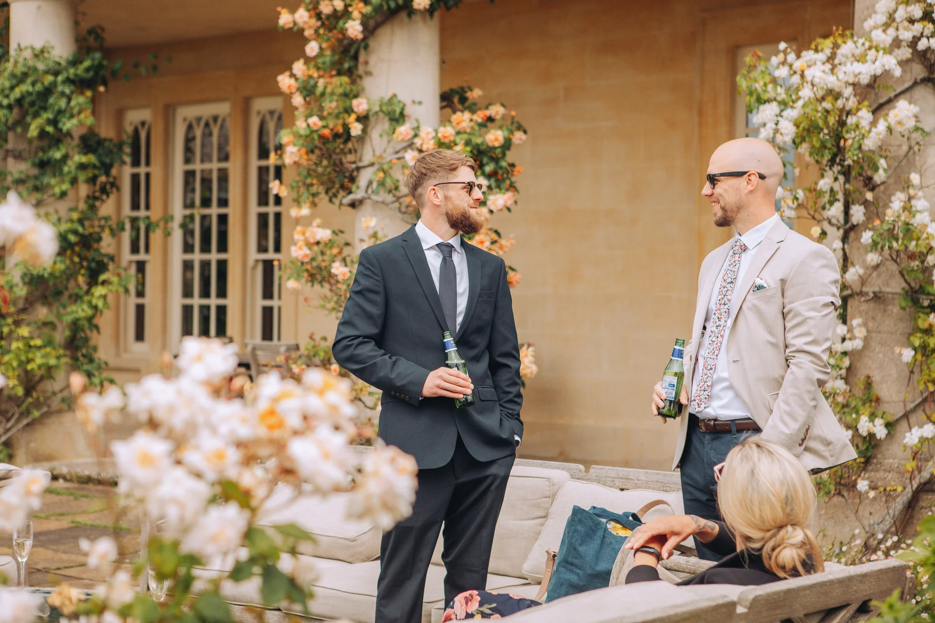 Two groomsmen wearing sunglasses and suits hold beer bottles and converse outdoors in front of a stone building decorated with climbing roses.