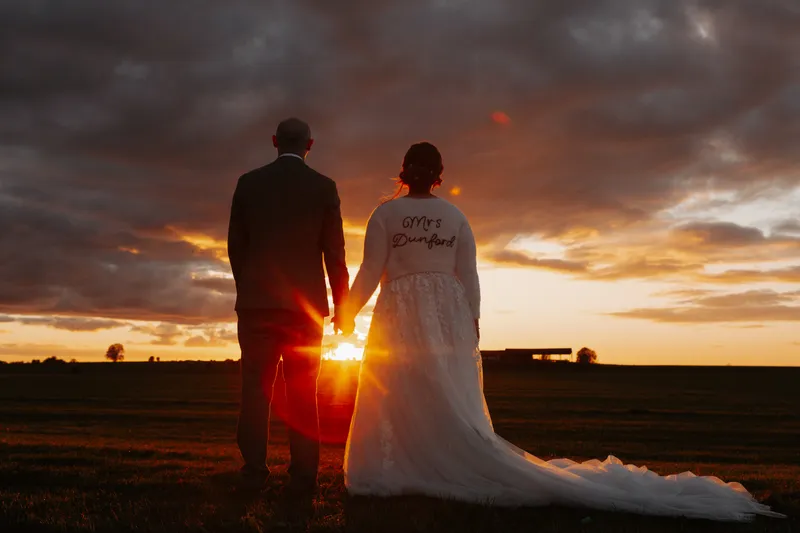 A newlywed couple holds hands while standing in a rural field, silhouetted against a dramatic sunset sky with the bride wearing a custom jacket.
