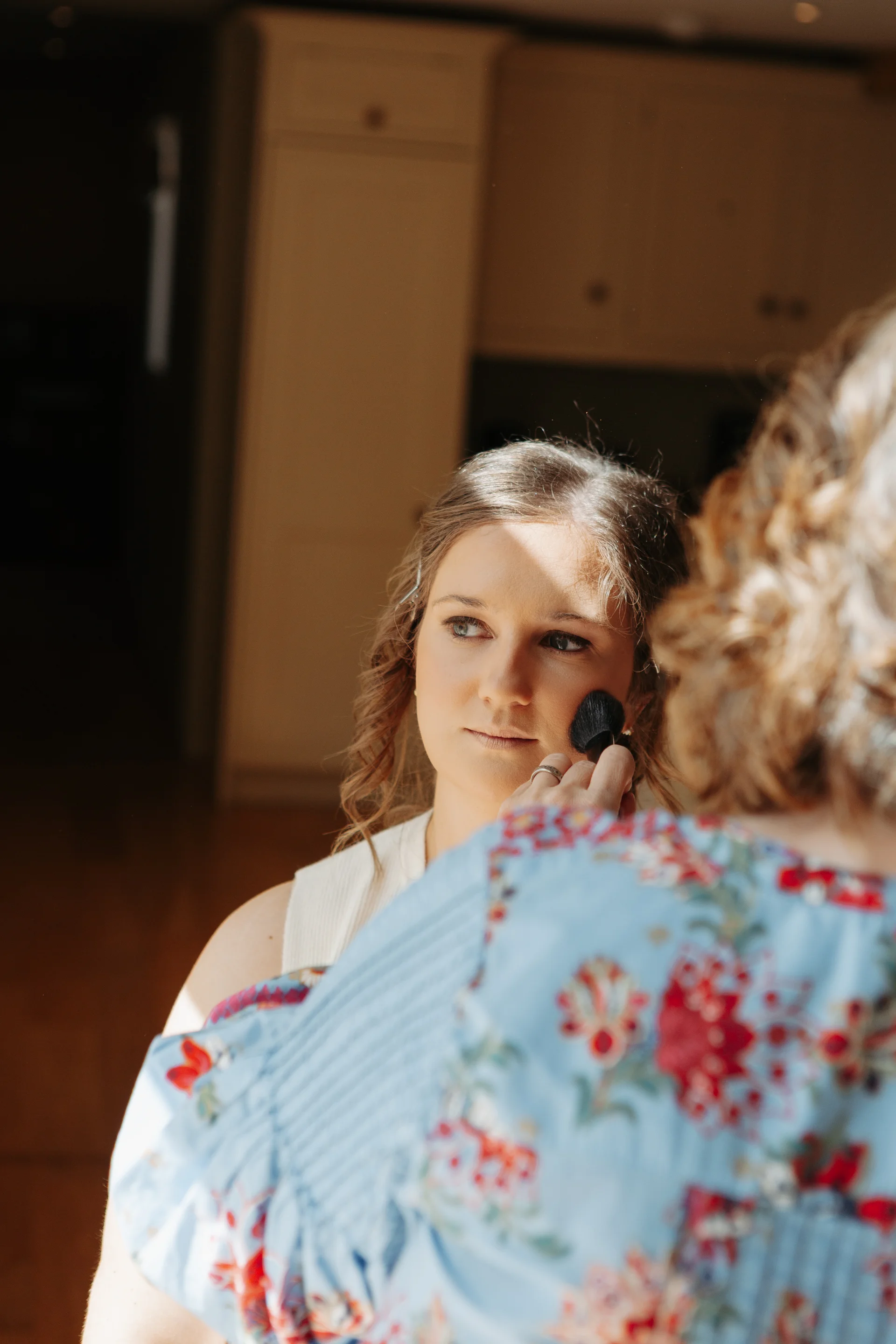 A bride having her makeup applied with a brush by a stylist during wedding morning preparations.