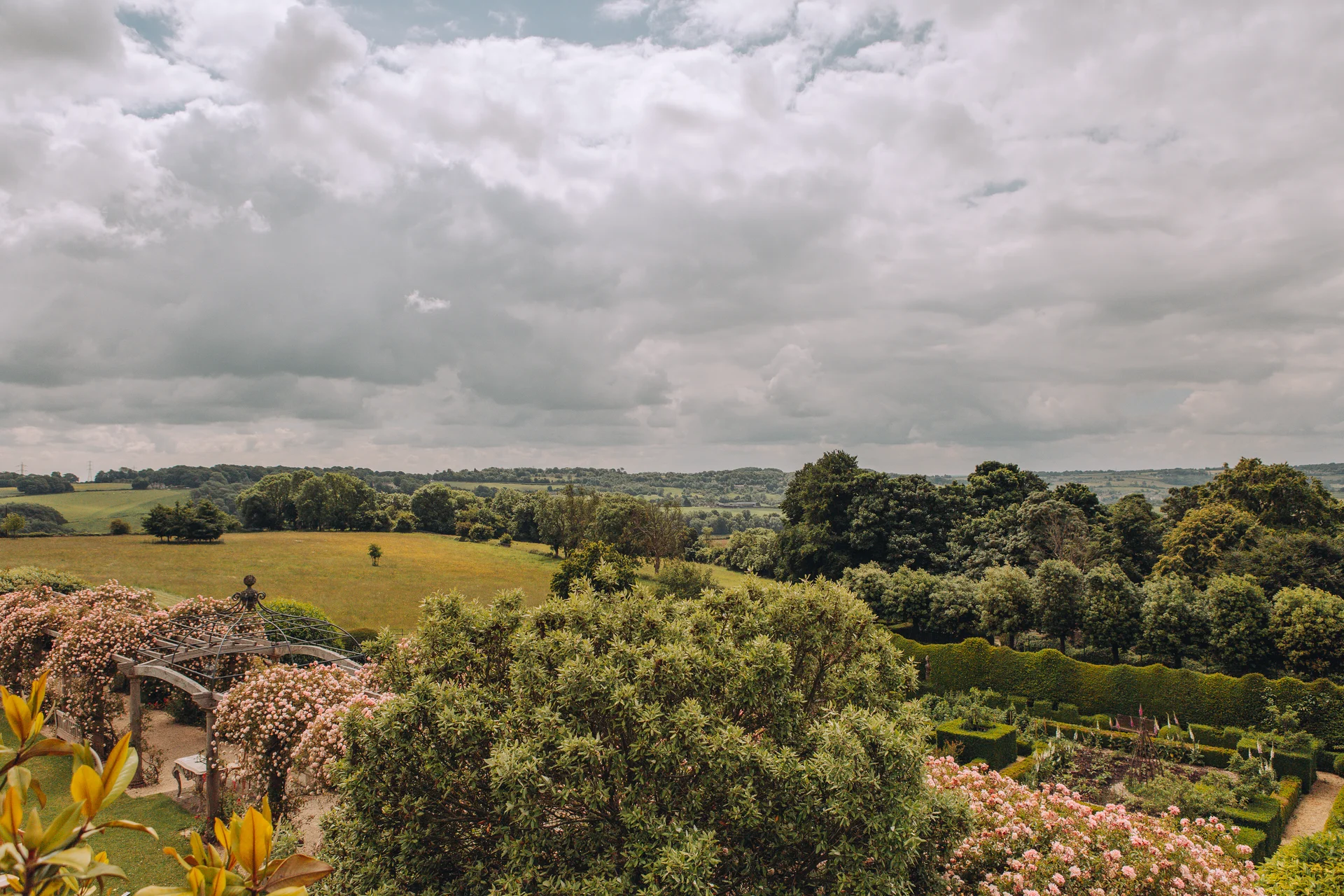 An expansive aerial view of an English country garden estate featuring a manicured walled garden, a wooden pergola, and a dramatic cloudy sky over rolling countryside.