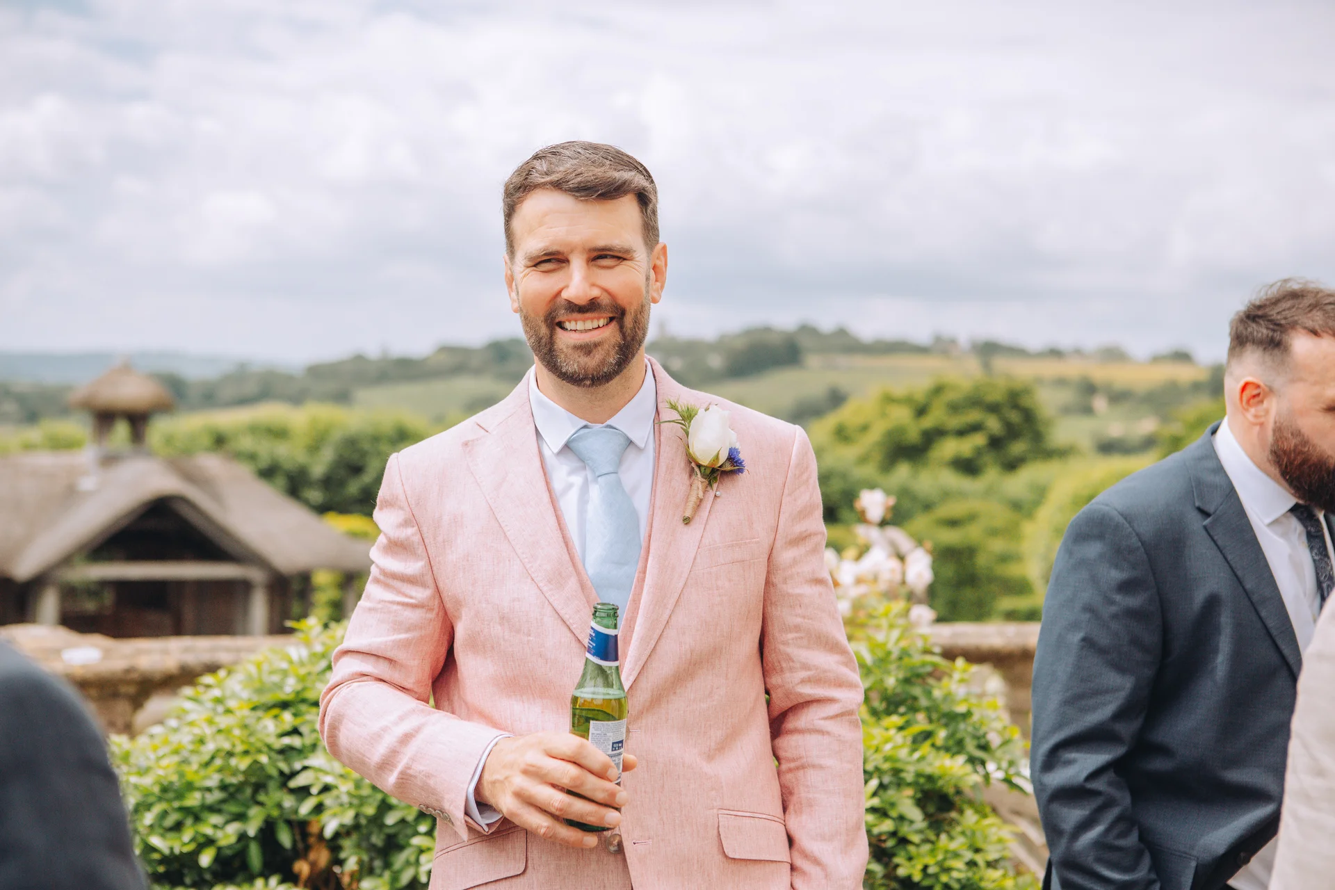 A smiling groom wearing a light pink suit jacket and pale blue tie holds a beer bottle outdoors during the wedding reception.