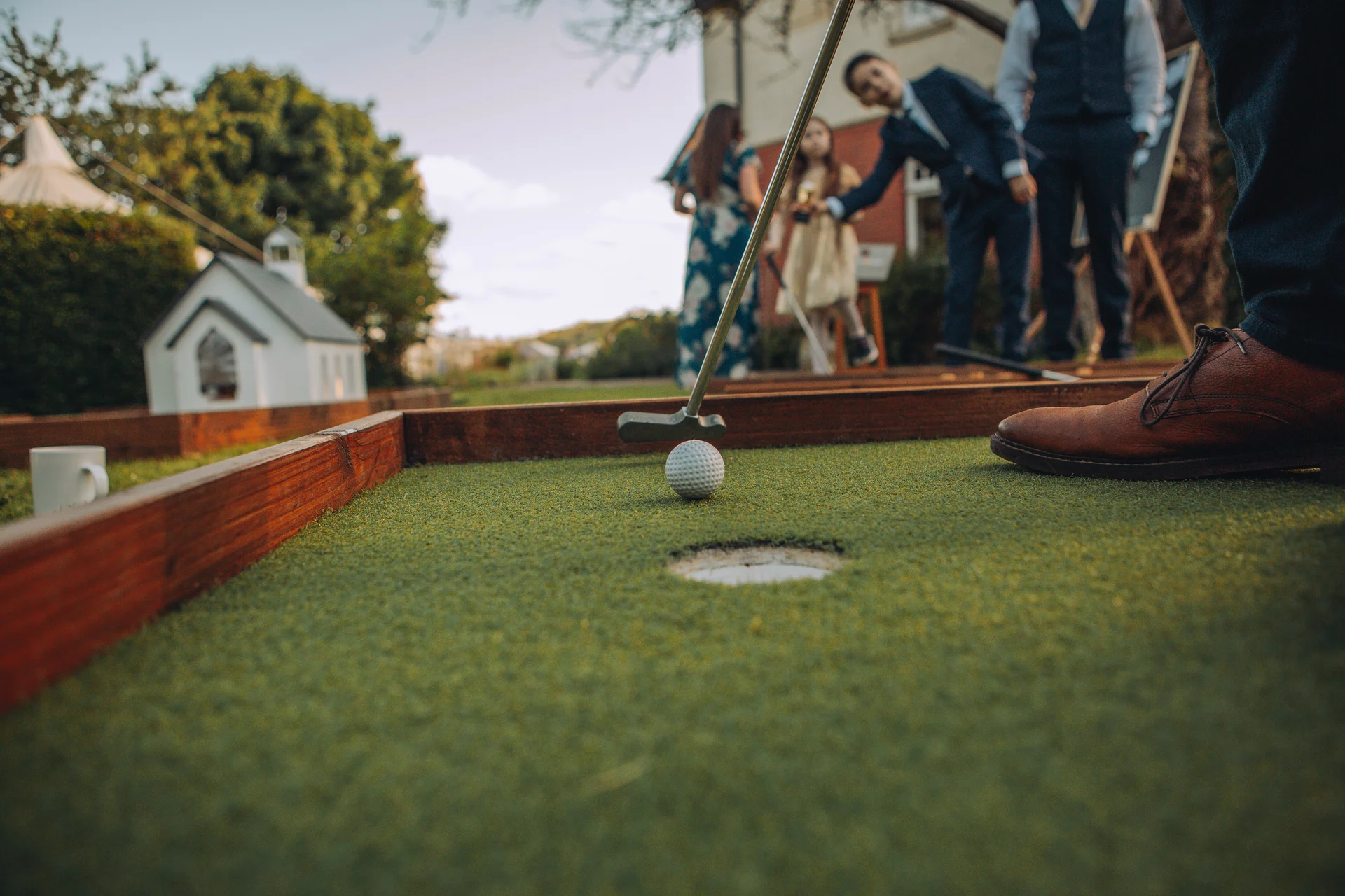 A low-angle close-up shot captures a guest playing crazy golf on a lawn during a wedding reception, with children watching in the background.