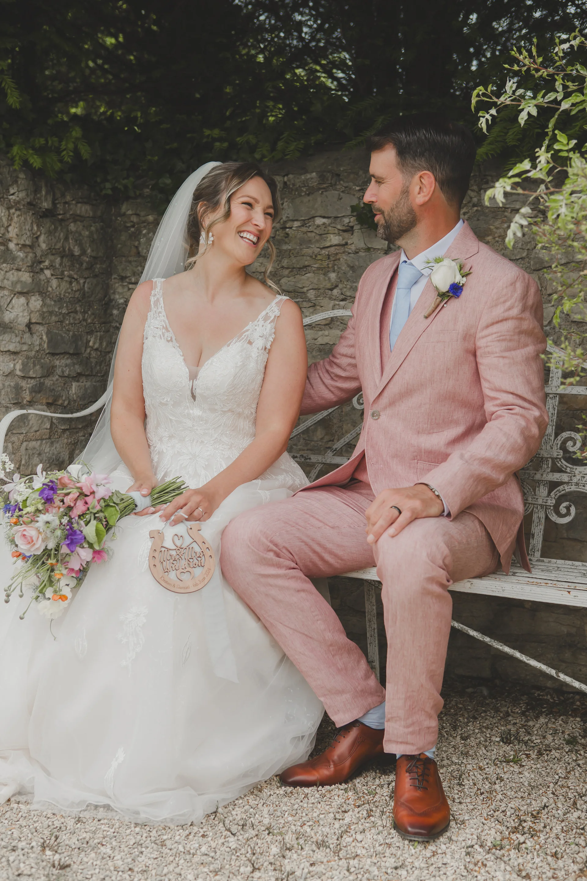 A newlywed couple laughing together while seated on an ornate white metal bench against a rustic stone wall, with the bride holding a colourful wildflower bouquet and a wooden horseshoe decoration.