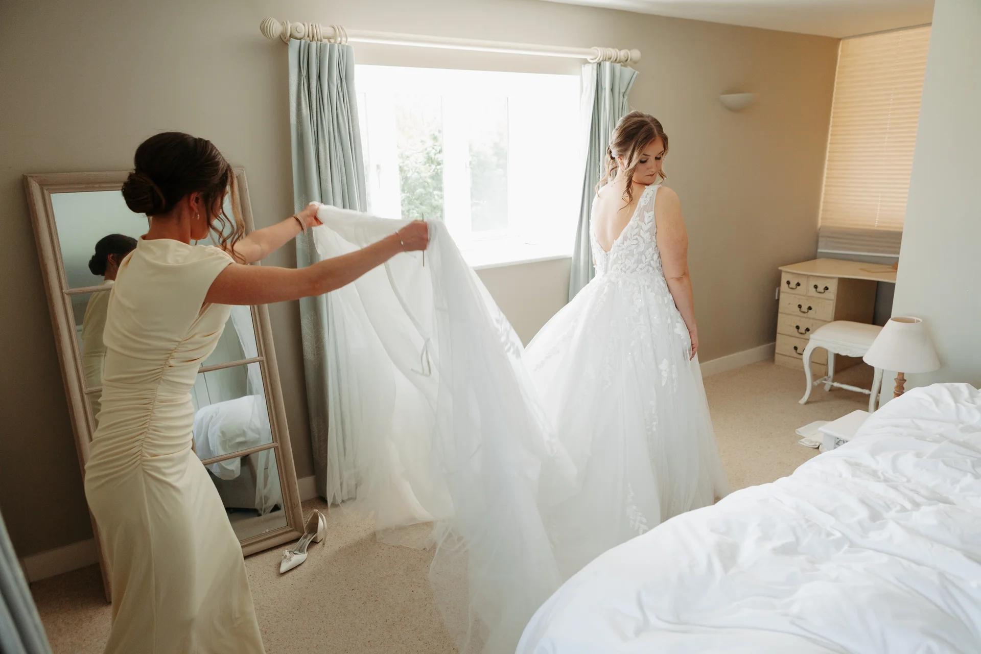 A bridesmaid holds up the flowing tulle train of a lace wedding gown as the bride looks back over her shoulder in a light-filled bedroom.