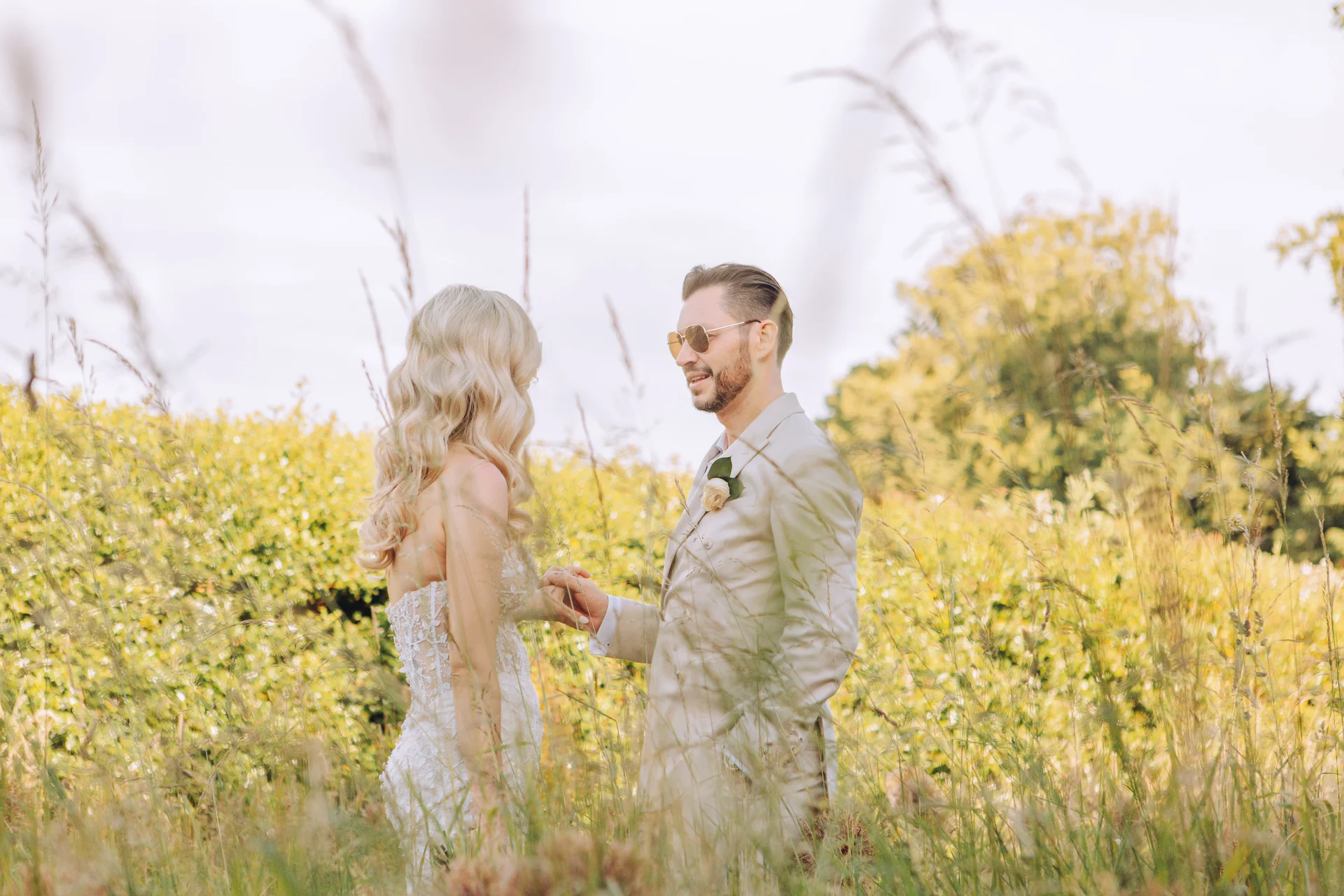 Newlyweds stand hand in hand within a sunlit wildflower meadow, framed by tall grass and foliage during an outdoor portrait session.