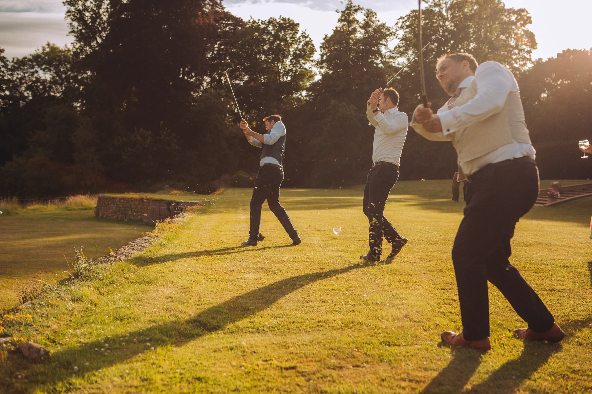 Three groomsmen play golf on a manicured lawn in the warm golden hour sunlight during an outdoor wedding reception.