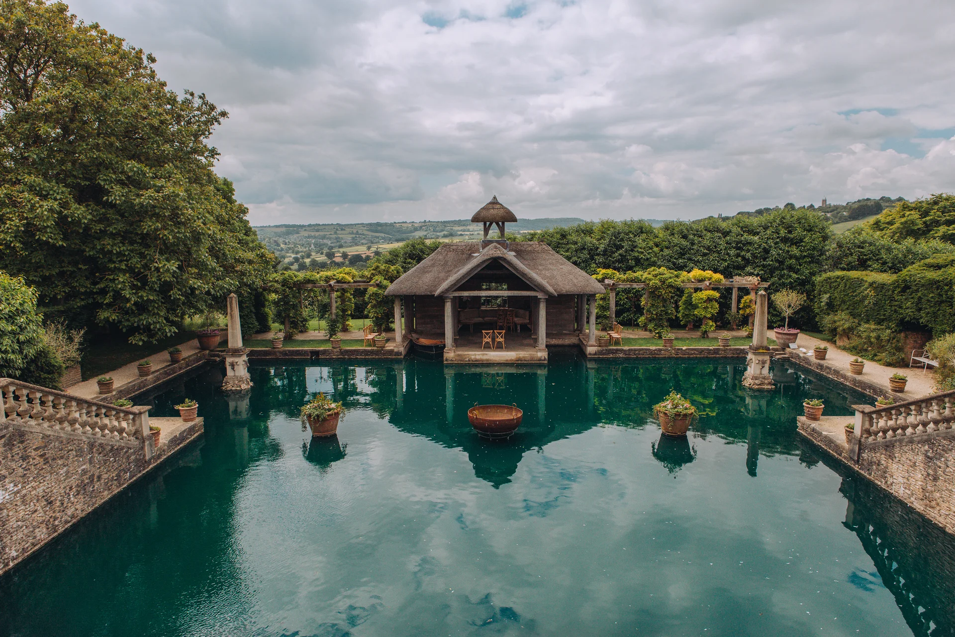 An expansive outdoor wedding venue featuring a stone-rimmed reflecting pool with a thatched pavilion at the far end, surrounded by lush trees and manicured gardens under a cloudy sky.
