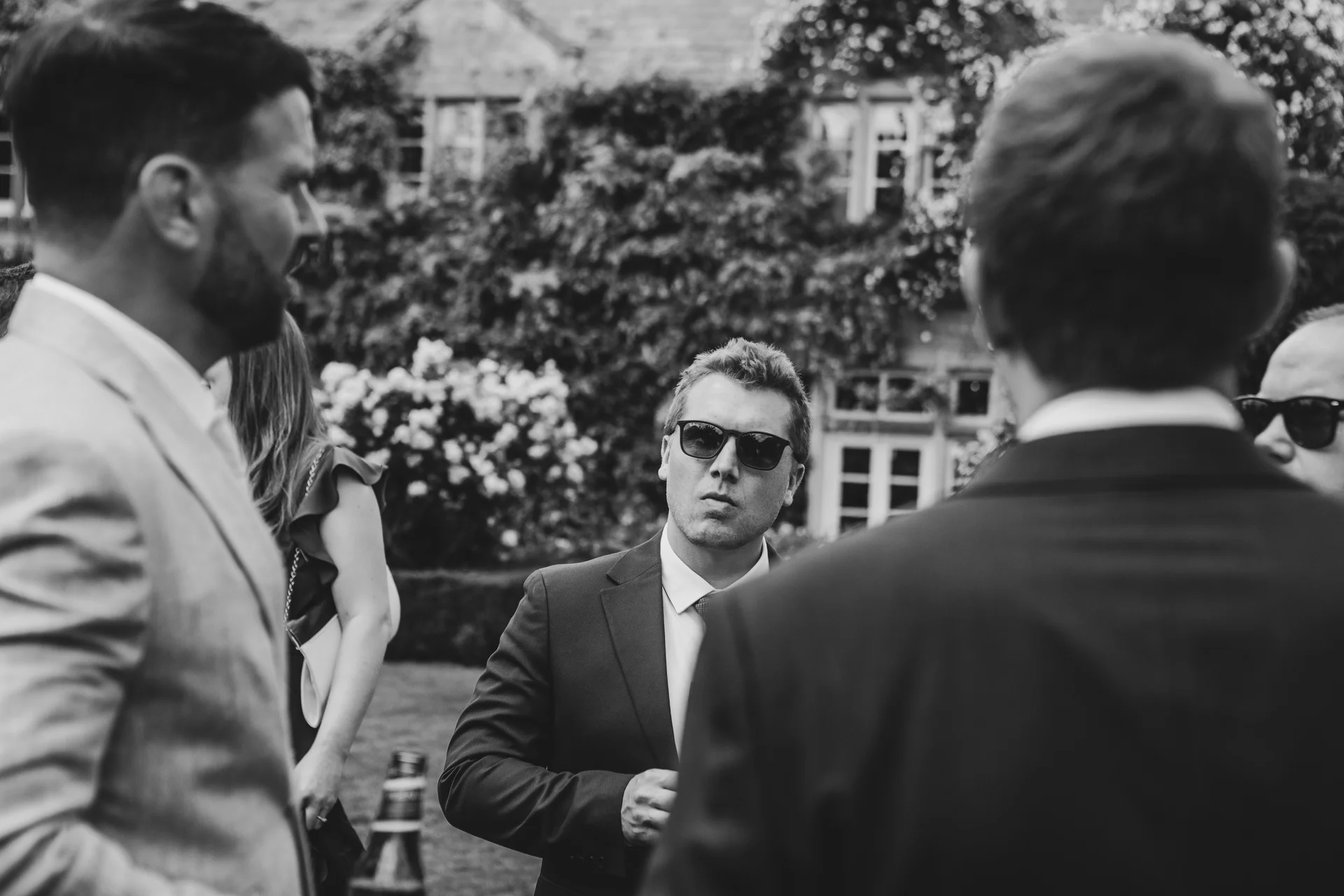 A candid black and white shot captures a group of wedding guests and groomsmen mingling outdoors in front of an ivy-clad manor house.