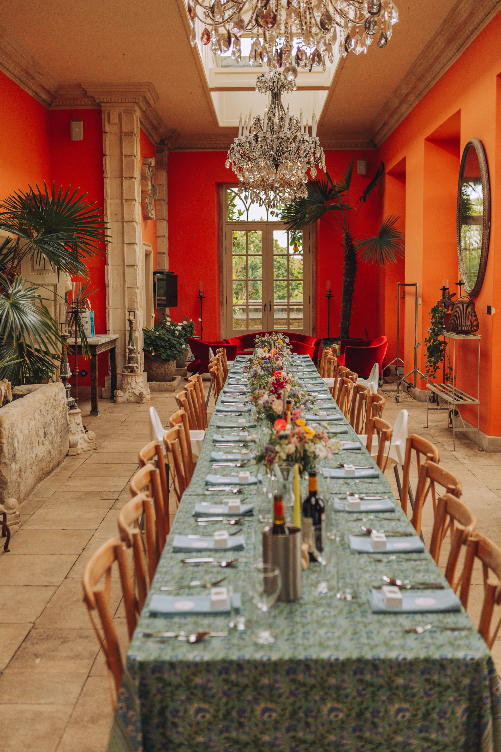 A long banquet table is elegantly set for a wedding reception inside a sunlit orangery featuring striking vibrant orange walls and crystal chandeliers.