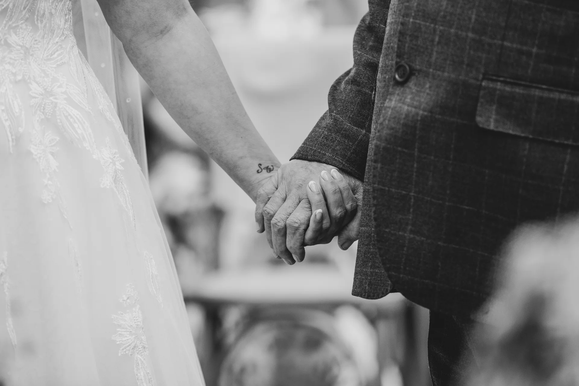 A close-up black and white photograph of a bride in a lace wedding dress holding hands with a groom wearing a checked suit jacket.