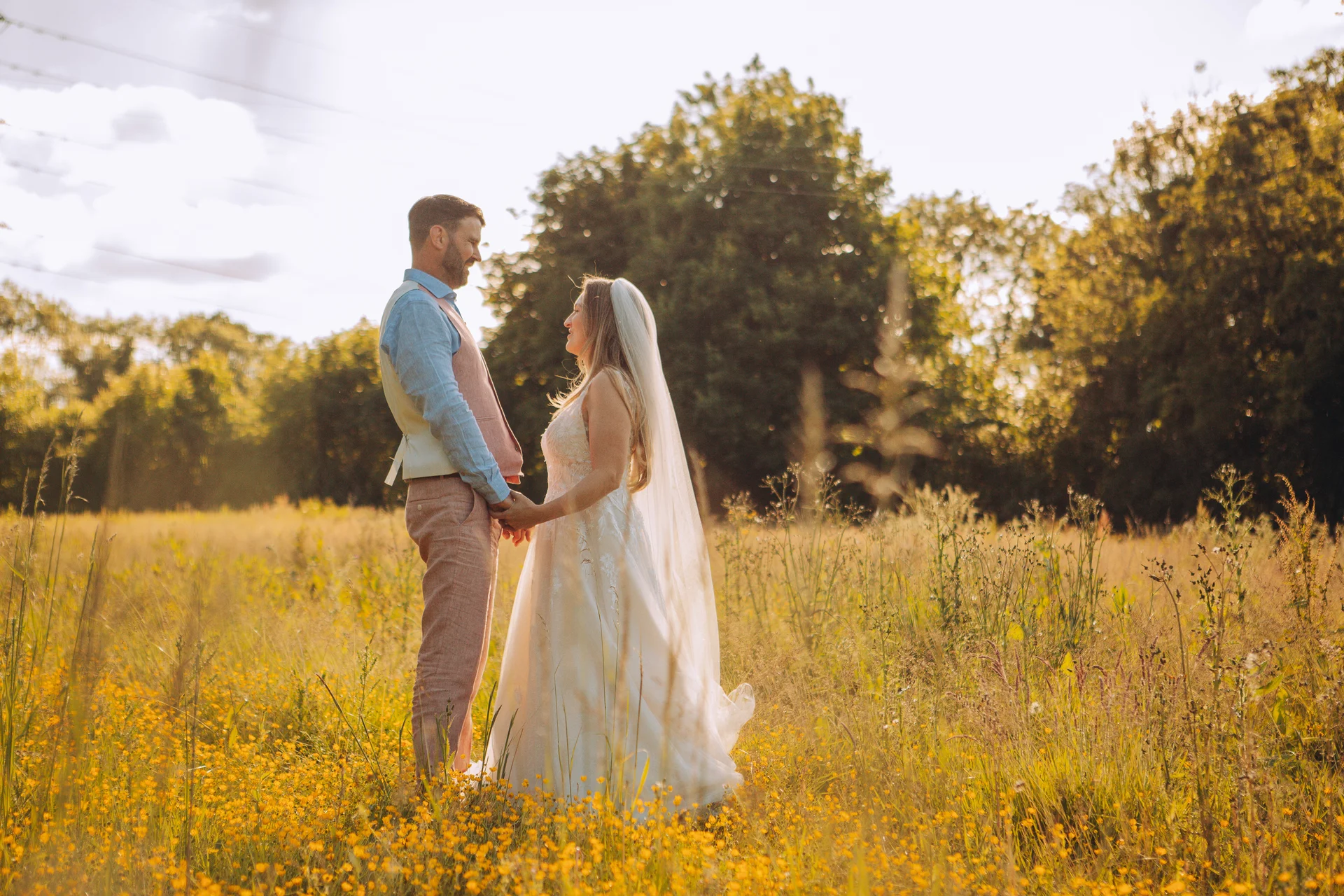 A newlywed couple stands hand-in-hand in a sunlit wildflower meadow during golden hour, bathed in warm backlighting.
