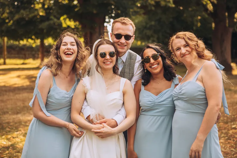 A joyful bride, groom and bridesmaids wearing sunglasses and pale blue dresses pose outdoors in the warm summer sunshine.