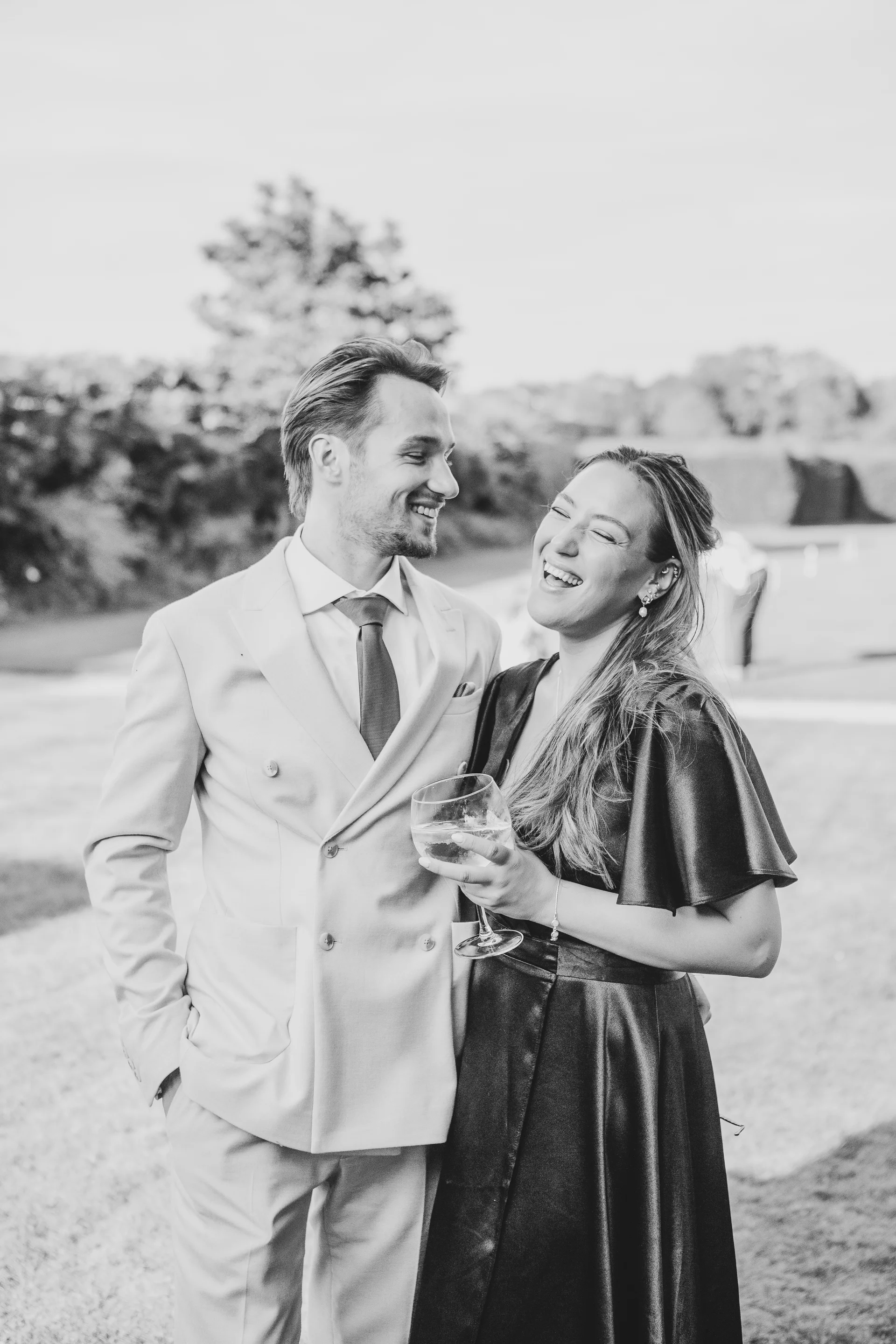 A stylish couple shares a joyful moment of laughter together outdoors during a wedding reception while holding a drink.