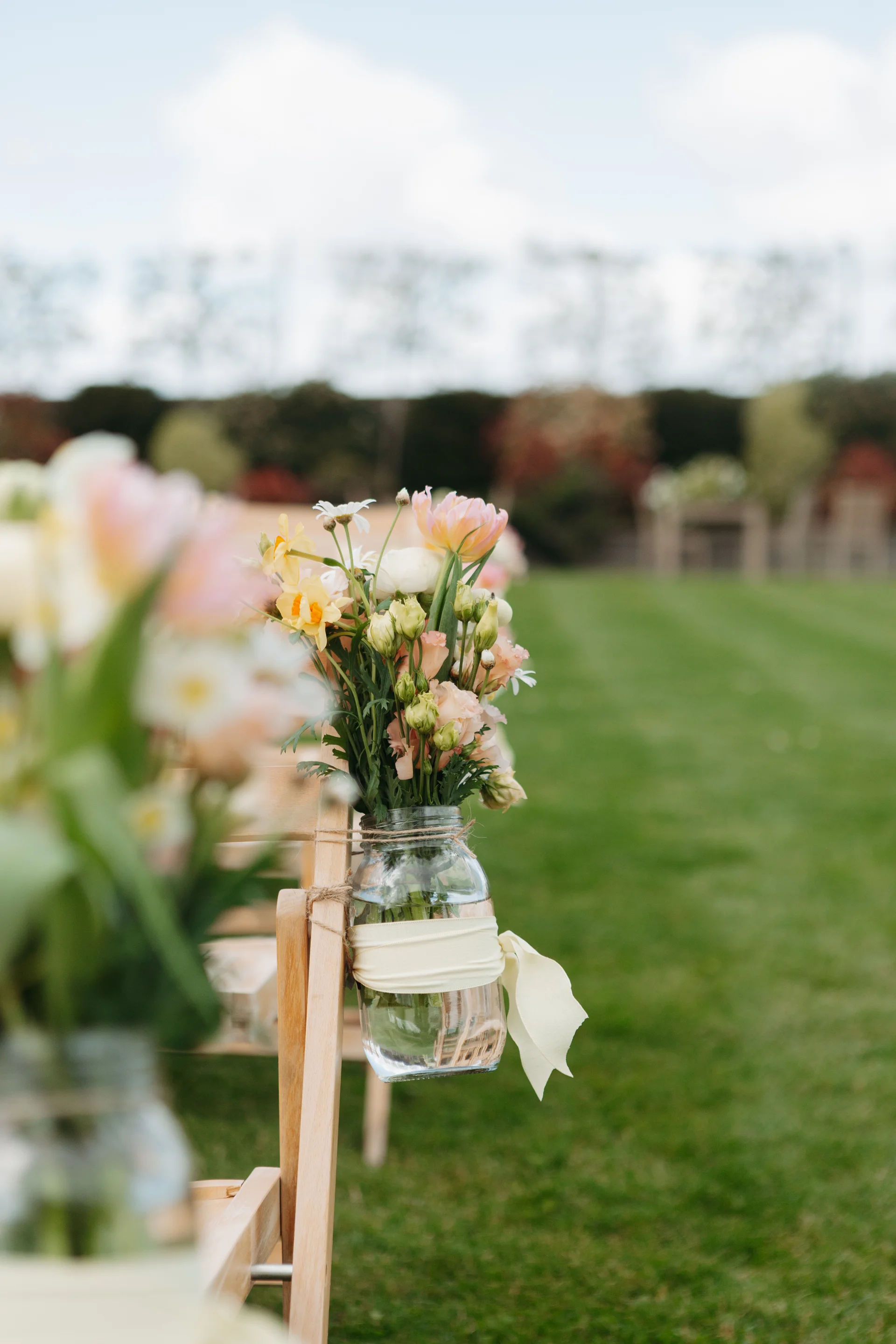 A close-up shot of a glass mason jar filled with pastel spring flowers and tied with a ribbon, hanging on a wooden chair during an outdoor wedding ceremony.