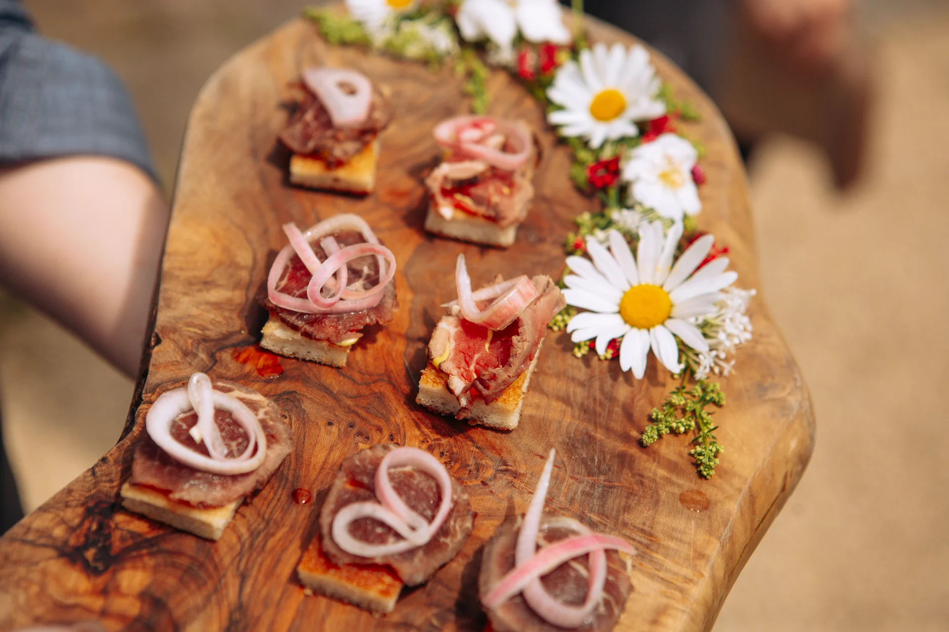 A rustic wooden board holds gourmet canapes garnished with pickled onion rings and decorated with fresh white daisies during an outdoor wedding drinks reception.