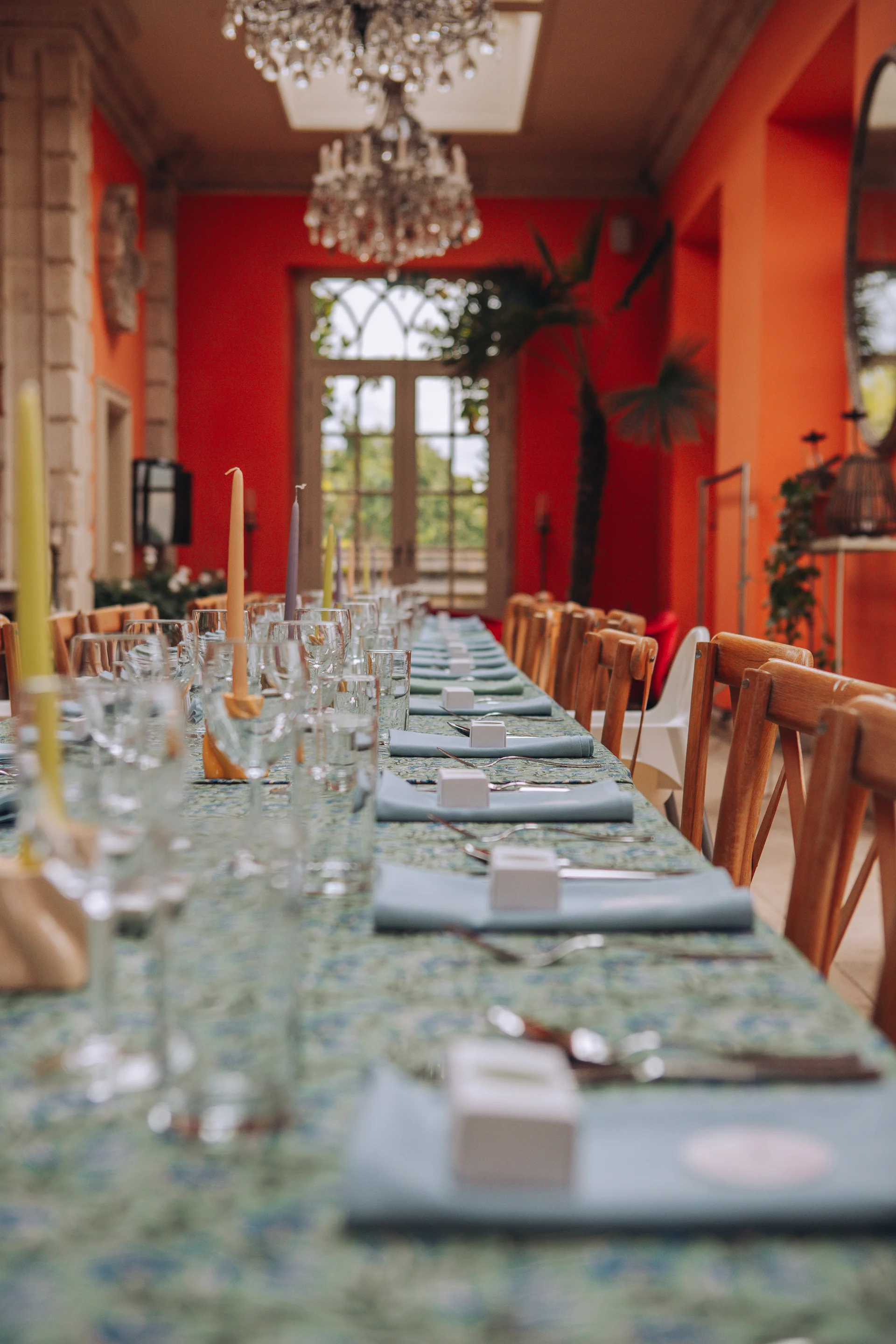 A long banquet table set for a wedding reception in a vibrant orangery featuring striking orange walls, patterned linens, and elegant taper candles.
