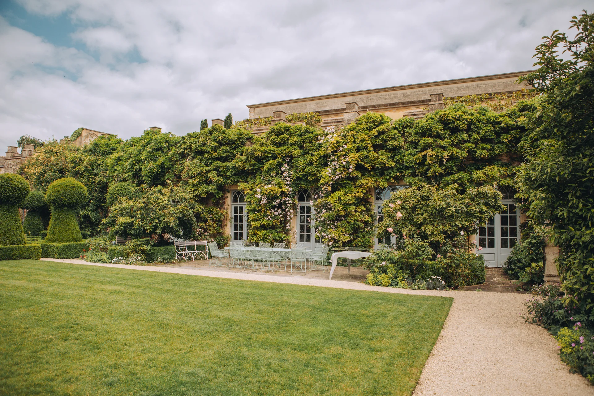 A stunning historic manor house featuring an ivy and climbing rose-covered exterior, elegant arched windows, and a manicured lawn leading to an outdoor terrace seating area.