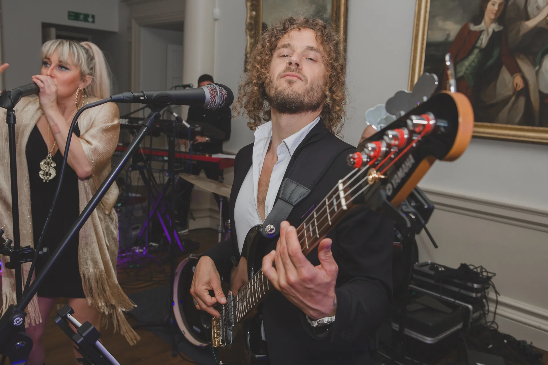 A charismatic bassist with curly hair plays his instrument passionately while a female singer performs alongside him in a lively wedding band.