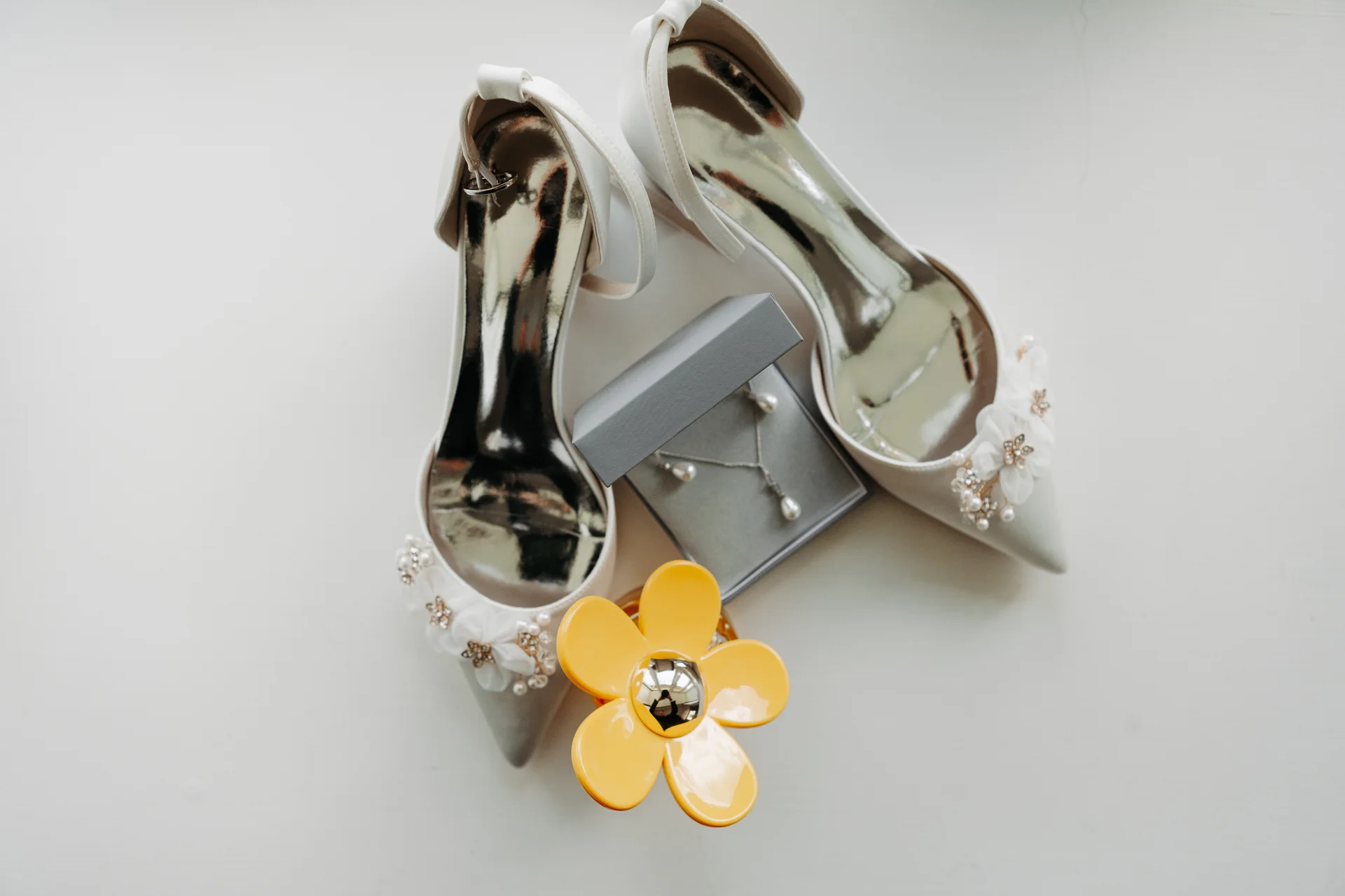An elegant bridal flat lay features a pair of satin heels with floral embellishments alongside a jewellery box and a yellow flower perfume bottle.