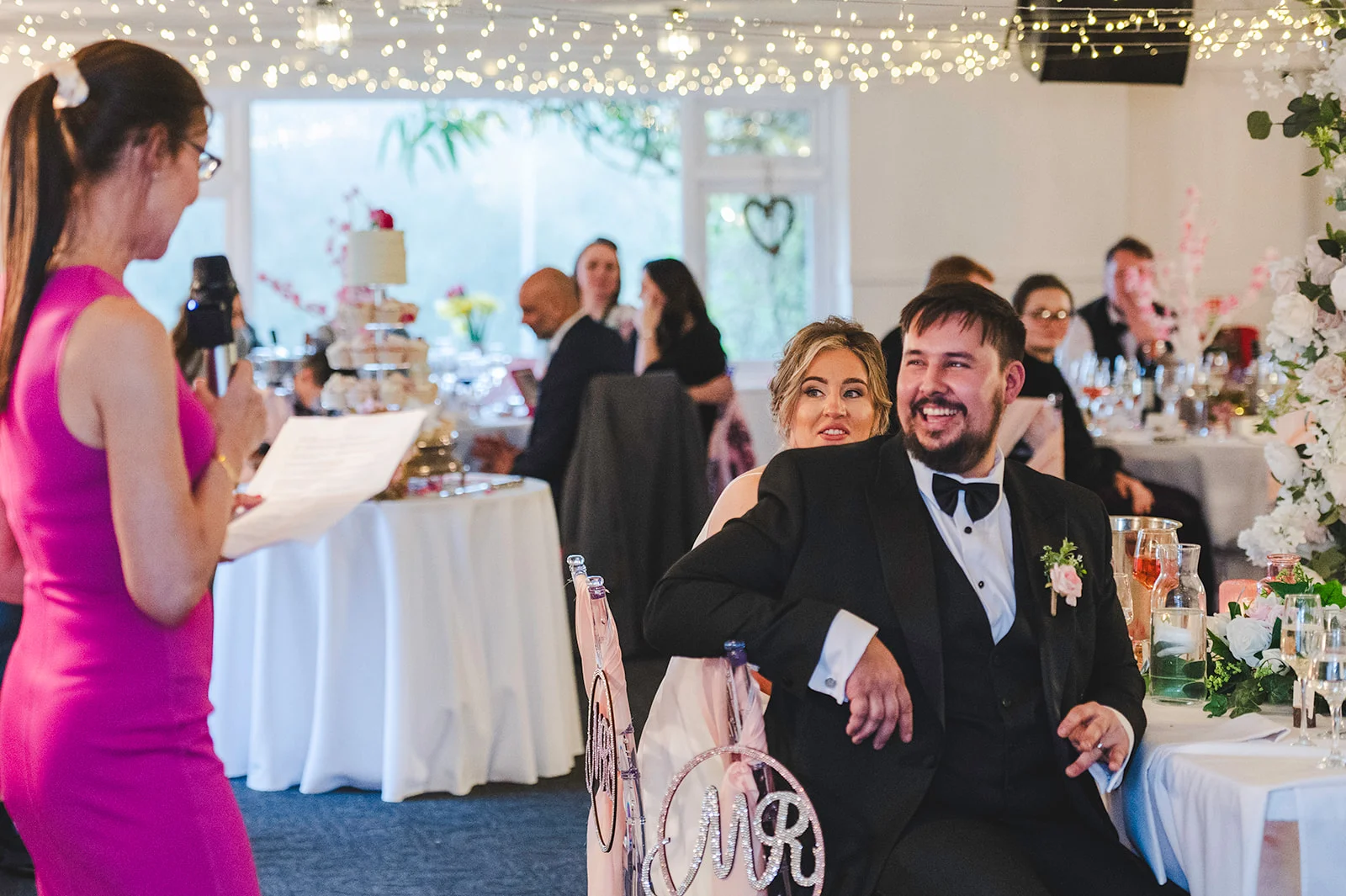 A joyful groom laughs heartily during a wedding speech at the top table, with the bride peeking over his shoulder under a canopy of fairy lights.
