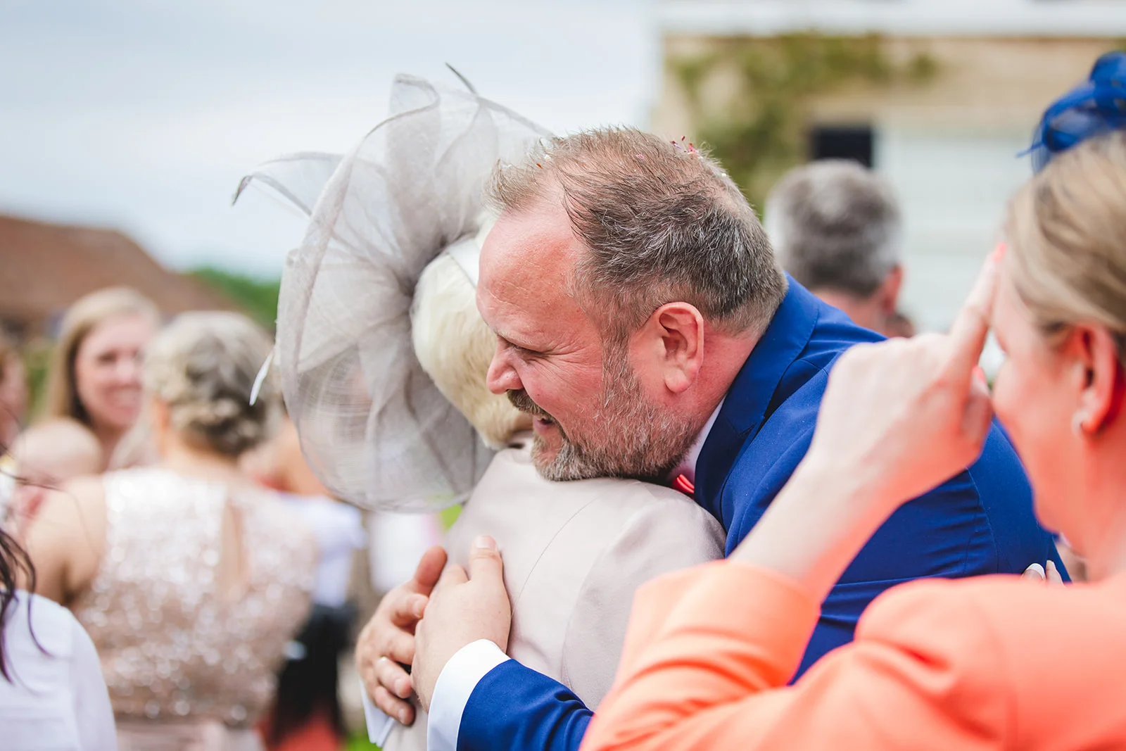 A guest in a blue suit warmly embraces another guest wearing a large fascinator during an outdoor wedding reception.