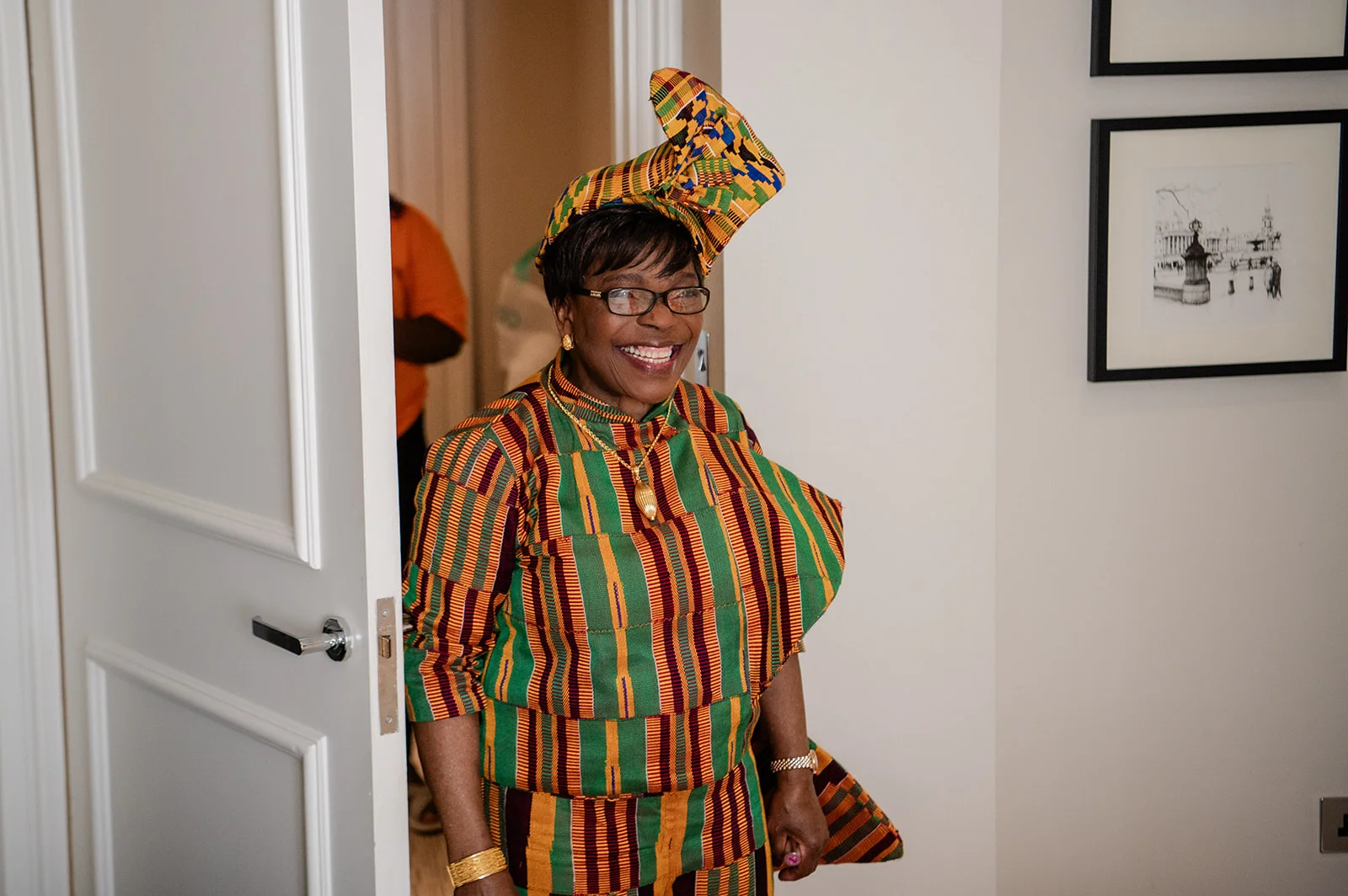 A smiling guest wearing a vibrant kente cloth outfit and matching headwrap stands inside a brightly lit room.