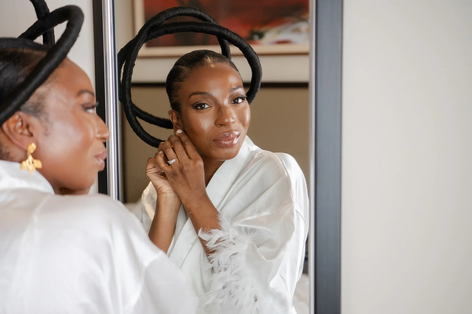 A smiling Black bride wearing a white robe with feather-trimmed sleeves puts on her earrings while looking into a mirror, showcasing a striking sculptural headpiece.