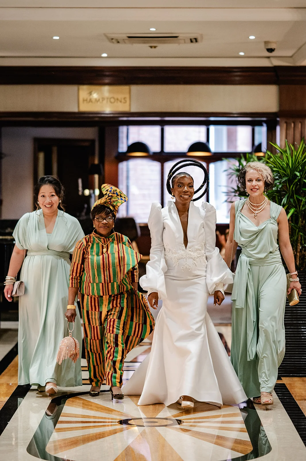 A smiling bride in a white gown and dramatic sculptural headpiece walks down a hotel hallway accompanied by her multi-ethnic bridal party in sage dresses and traditional patterned attire.
