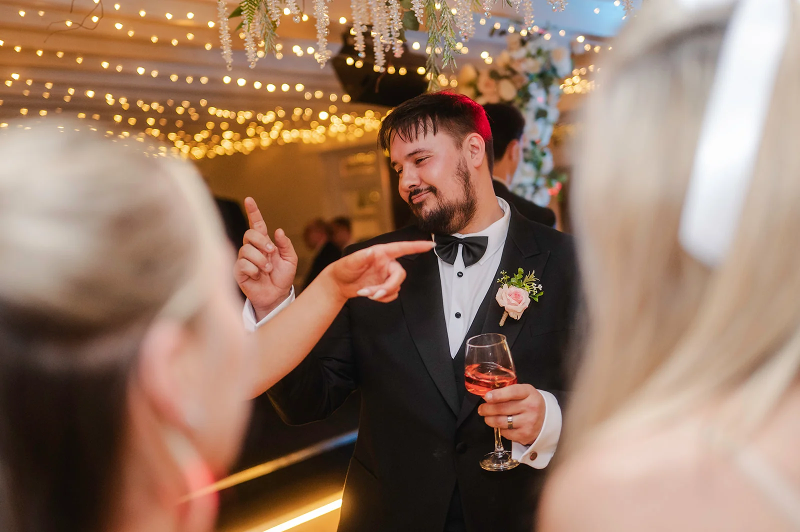 A groom in a black tuxedo with a pink rose boutonniere smiles and holds a glass of wine while mingling under a canopy of fairy lights during the wedding reception.