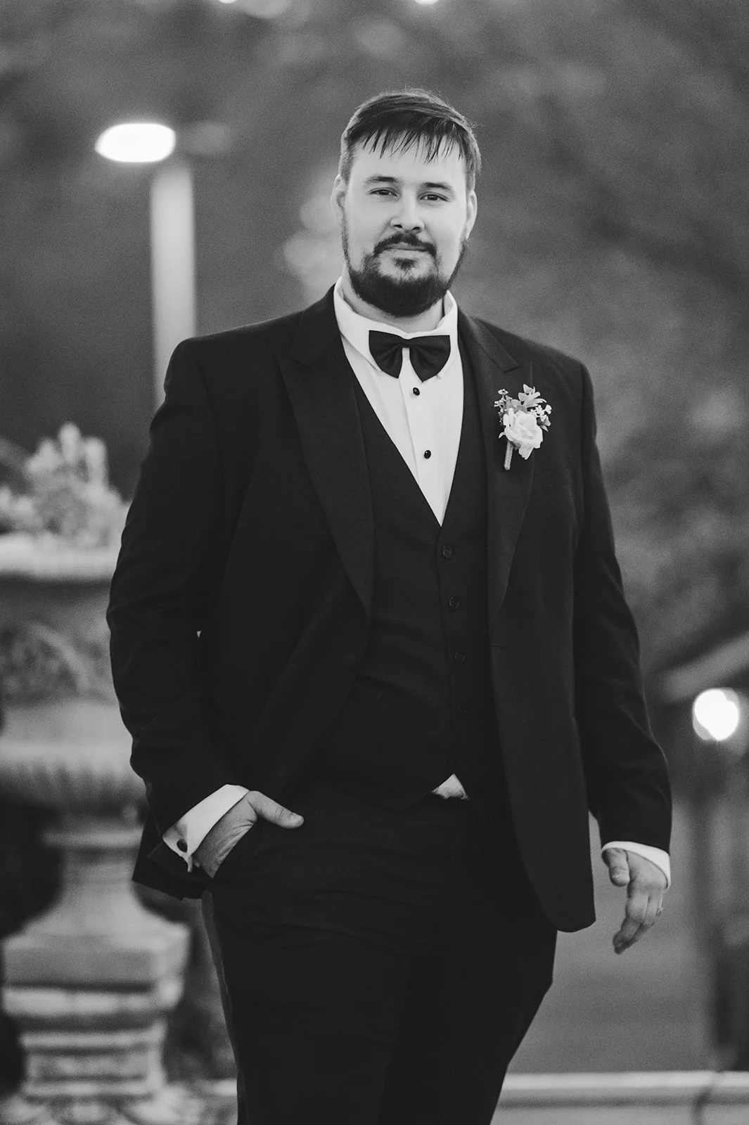 A handsome groom in a black tuxedo and bow tie poses confidently with one hand in his pocket during an evening wedding portrait.