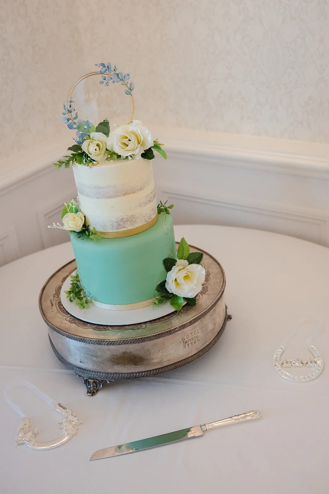A elegant two-tiered wedding cake featuring a sage green bottom tier and a semi-naked top tier decorated with white roses and an acrylic hoop topper.