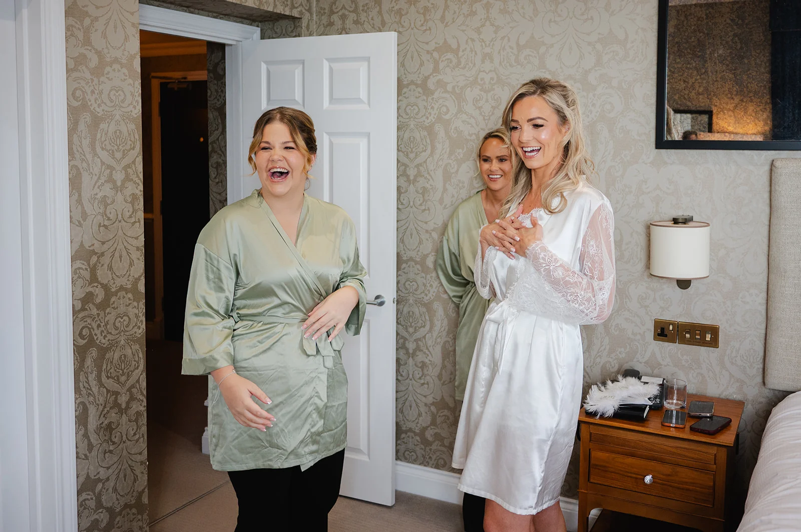 A joyful bride in a white lace-sleeved robe laughs heartily alongside bridesmaids wearing matching sage green robes during morning bridal preparations.
