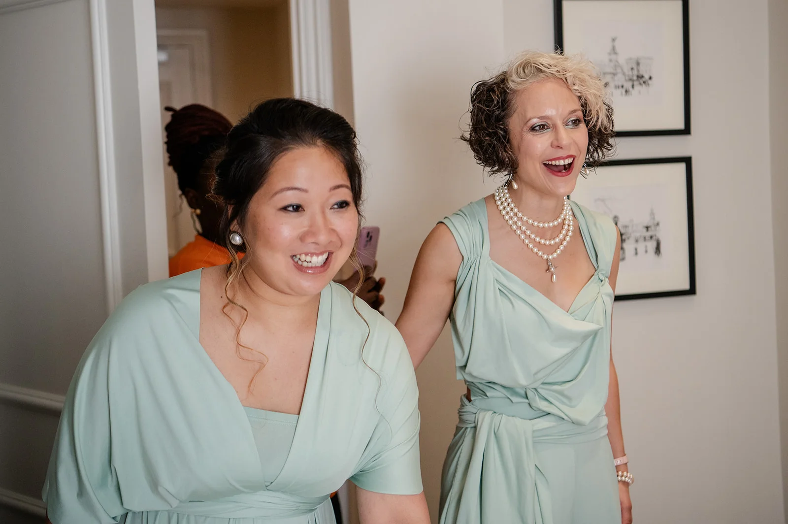 Two smiling bridesmaids wearing matching sage green dresses and pearl jewellery laugh happily while getting ready indoors.
