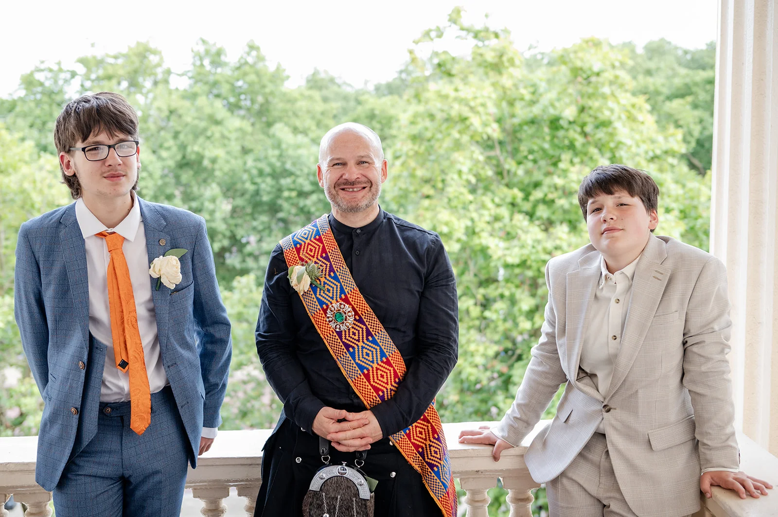 A smiling groom wearing a traditional kilt and colourful tartan sash stands outdoors on a balcony flanked by two groomsmen in smart suits.