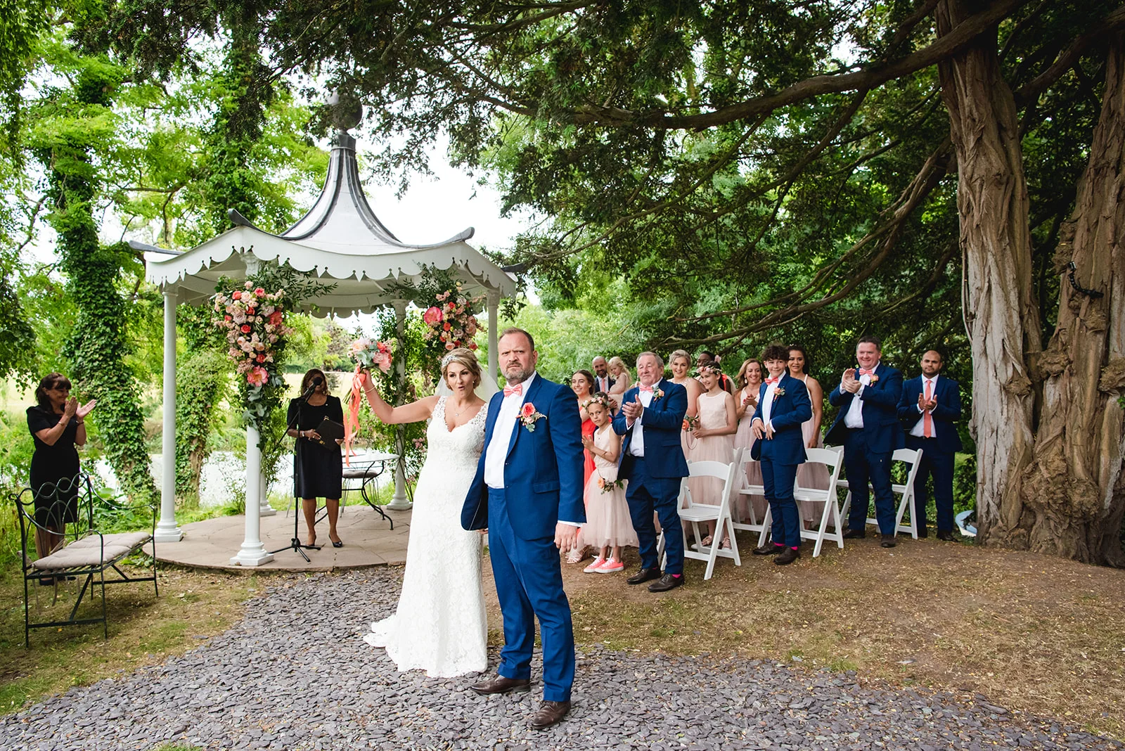 A newly married couple stands proudly on a cobbled path after an outdoor ceremony by a white bandstand, with guests clapping in the background under large trees.